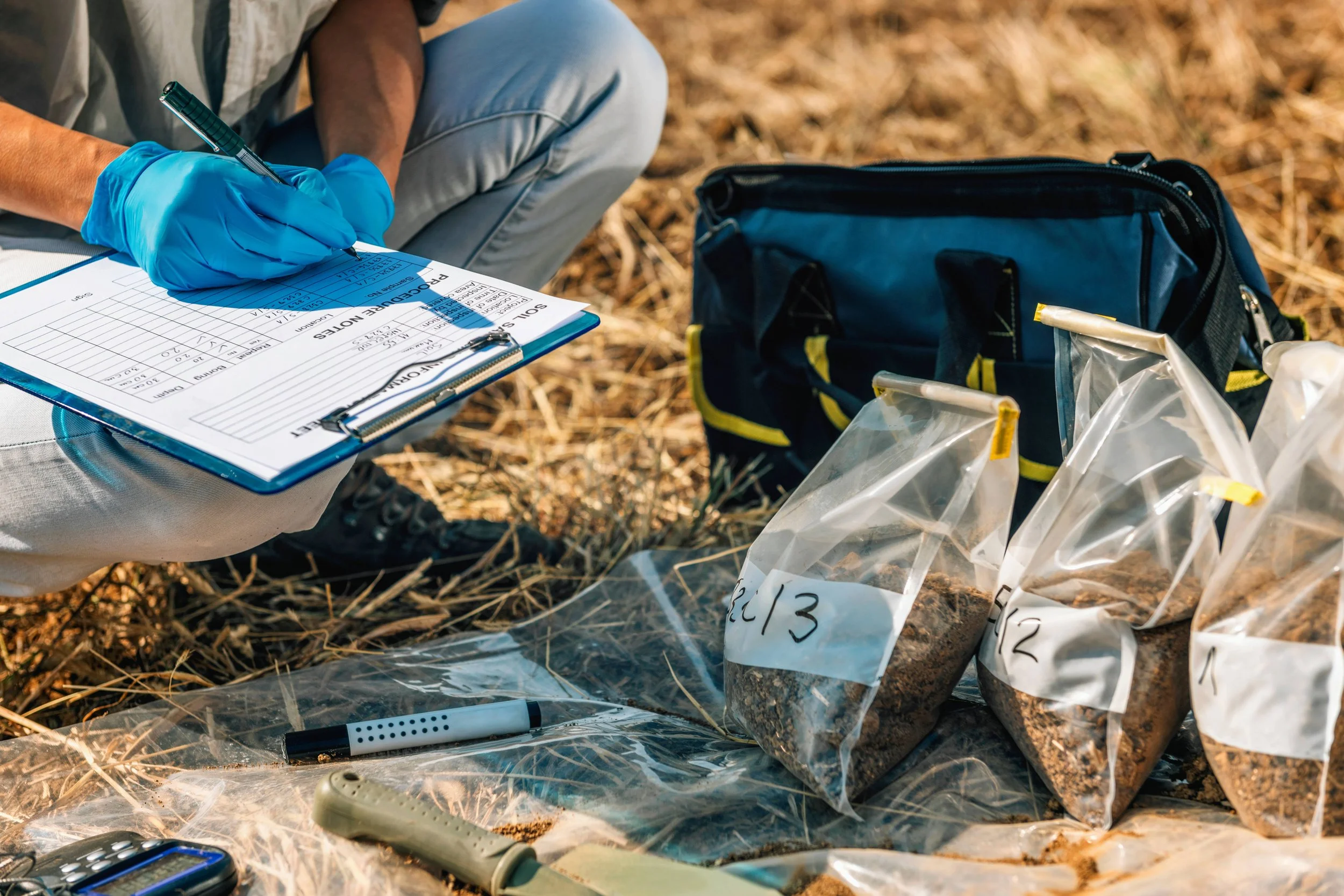 Person in blue gloves writing on a clipboard with medical or research data, surrounded by lab supplies, bags with samples, and outdoor environment with dry grass.