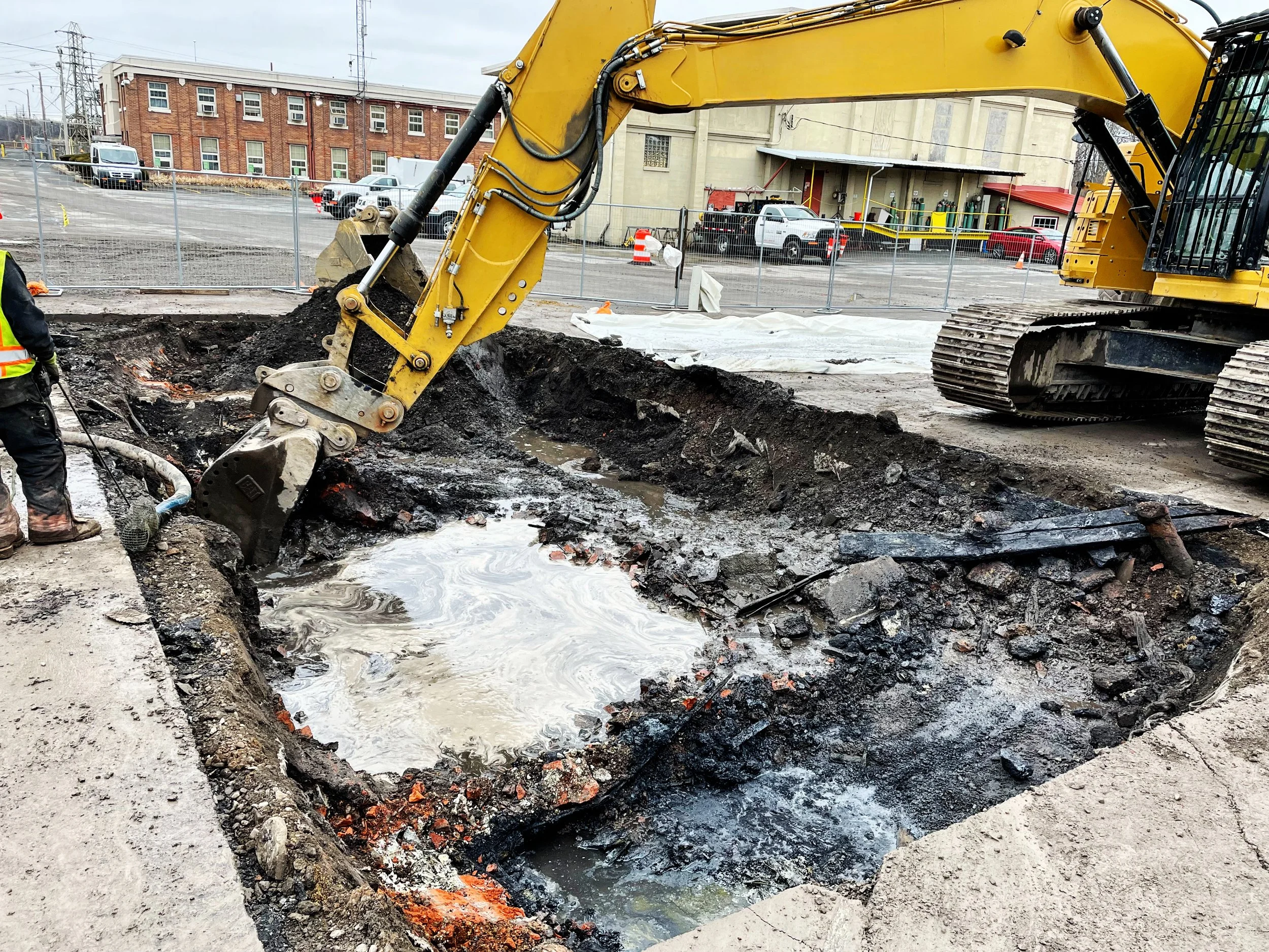 Construction site with a yellow excavator digging a hole in the ground, with water and debris, surrounded by a metal fence, and a background of parking lot and a large brick building.