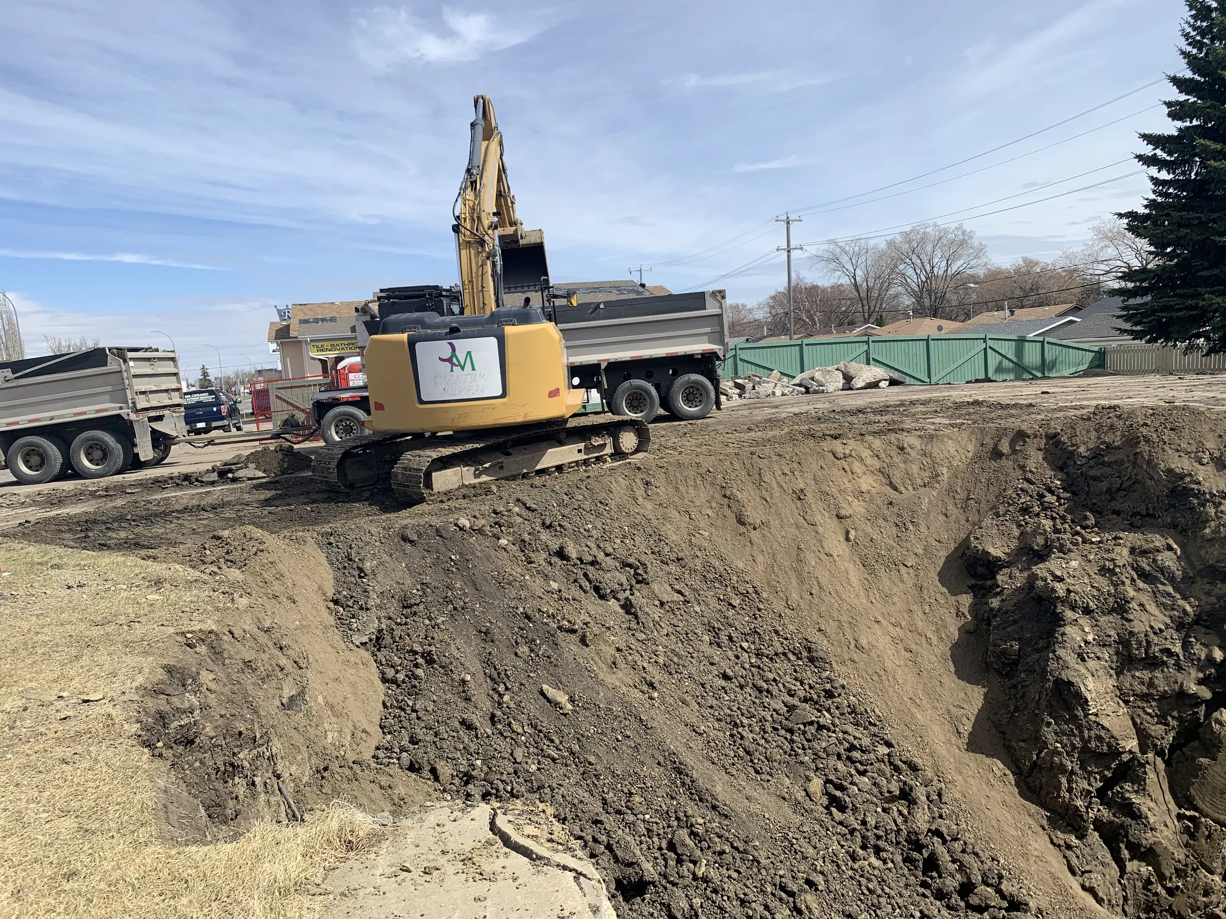 Construction site with a yellow excavator loading dirt into a dump truck. In the background, there are houses, a green fence, and utility poles under a partly cloudy sky.