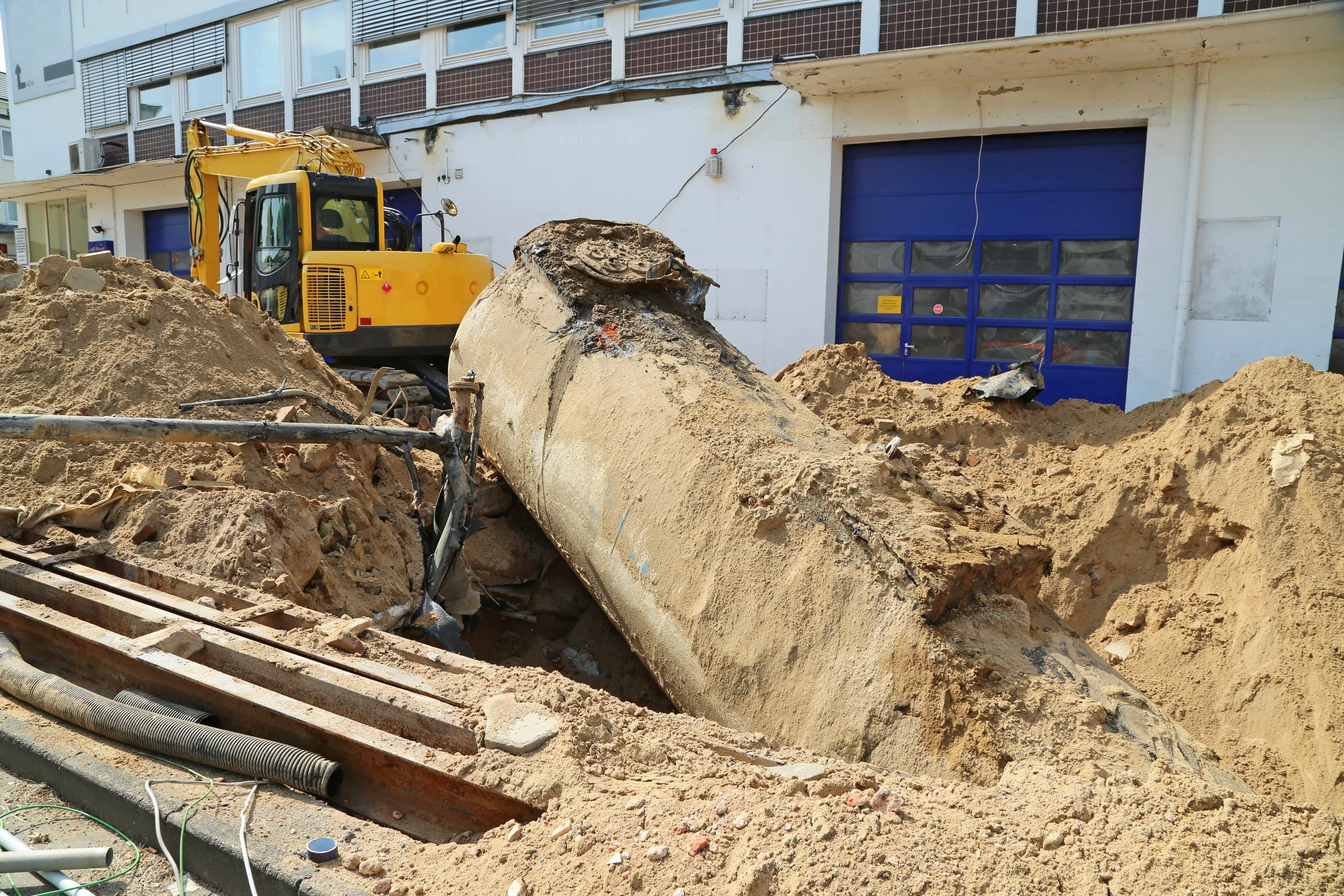 Construction site with a large pipe in the foreground surrounded by dirt and debris, with a yellow excavator and building with blue doors in the background.