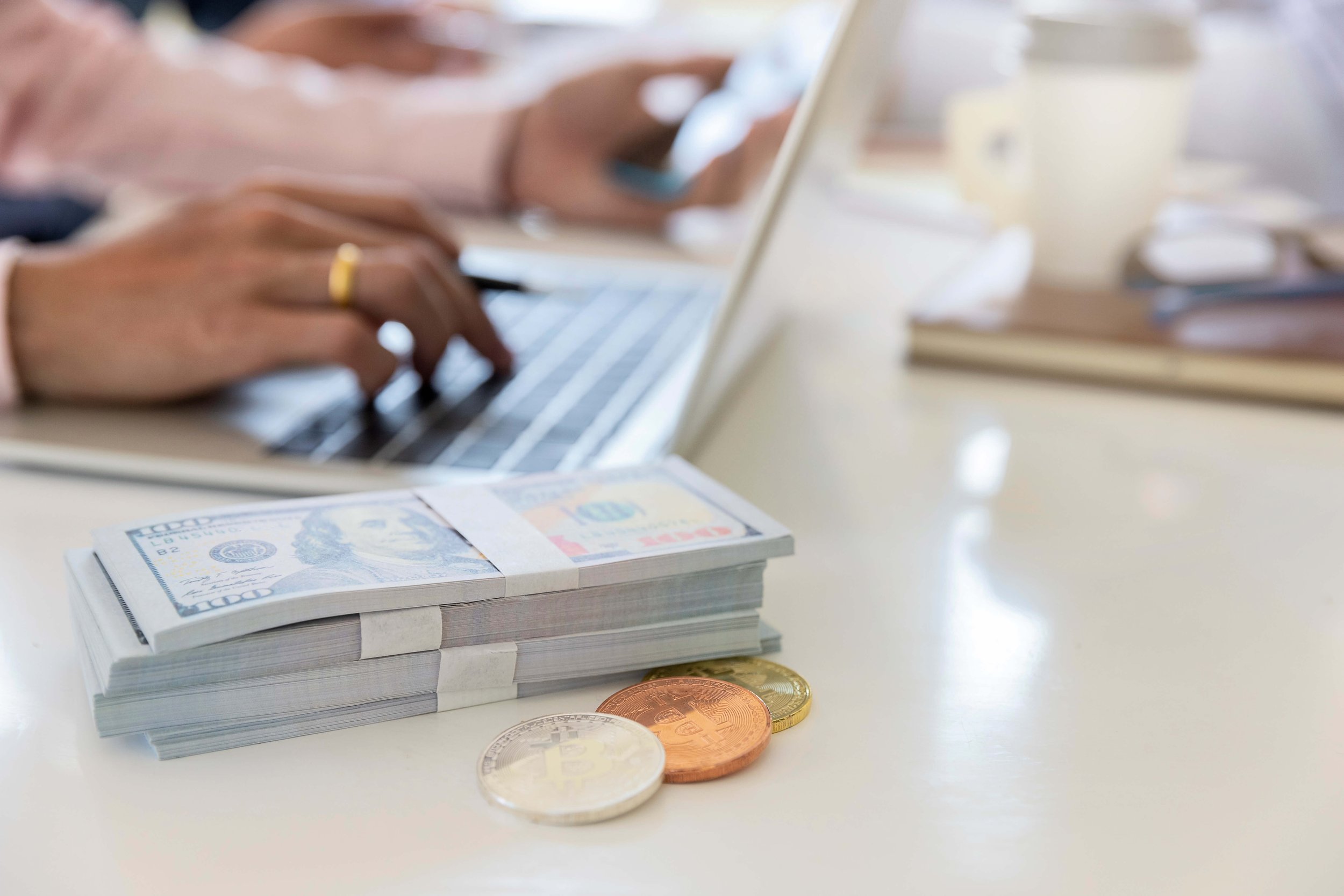 Stacks of US dollar bills and cryptocurrency coins on a table with someone using a laptop in the background.