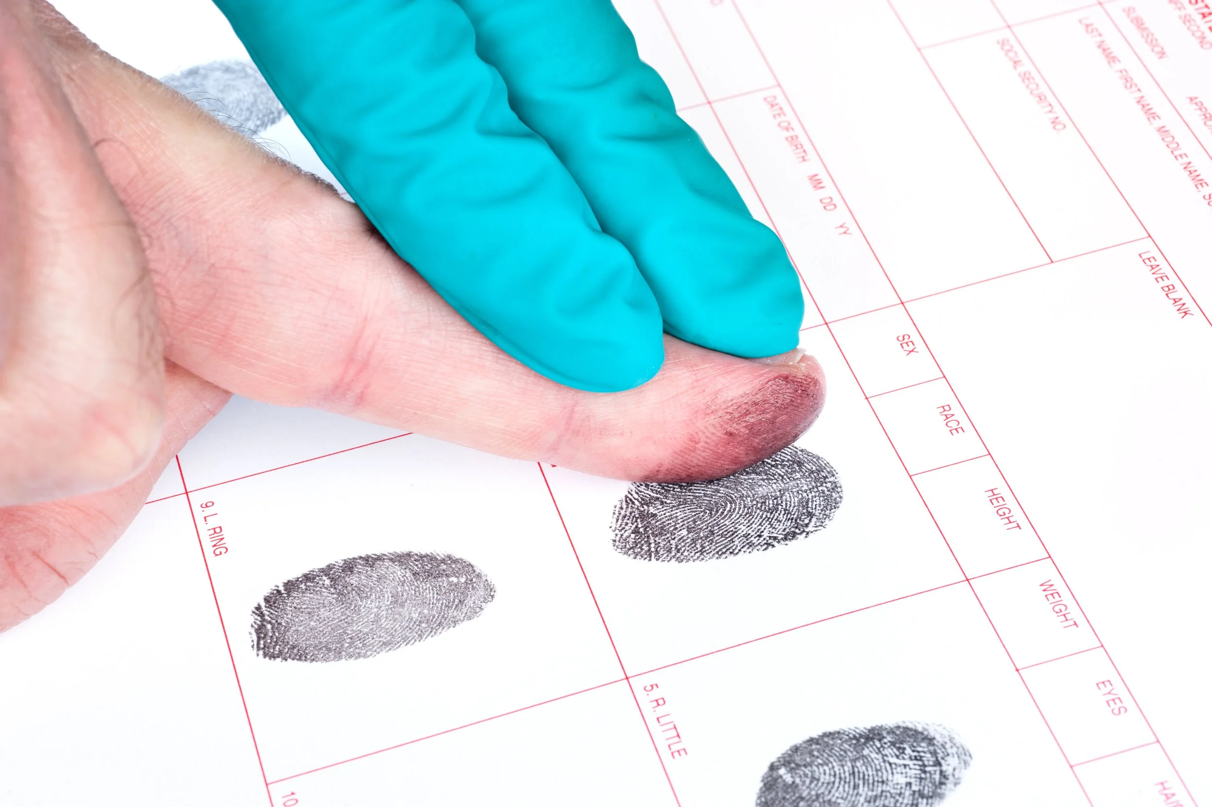 A finger with ink fingerprint on a sheet of paper, with fingers being examined by someone wearing a blue glove, showing fingerprint details for identification.