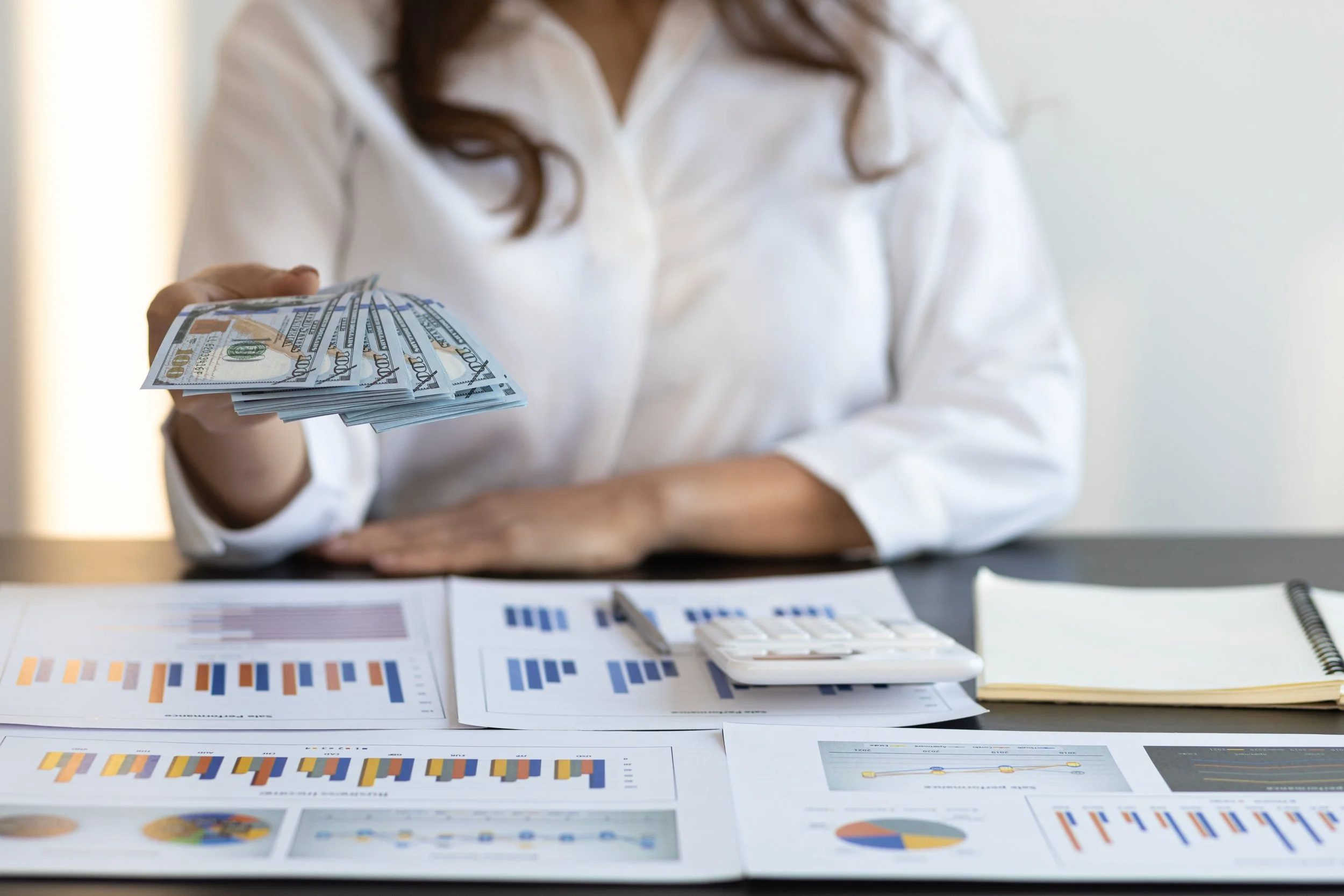 Person holding a fan of hundred-dollar bills on a desk with financial charts, documents, a calculator, and a notebook.