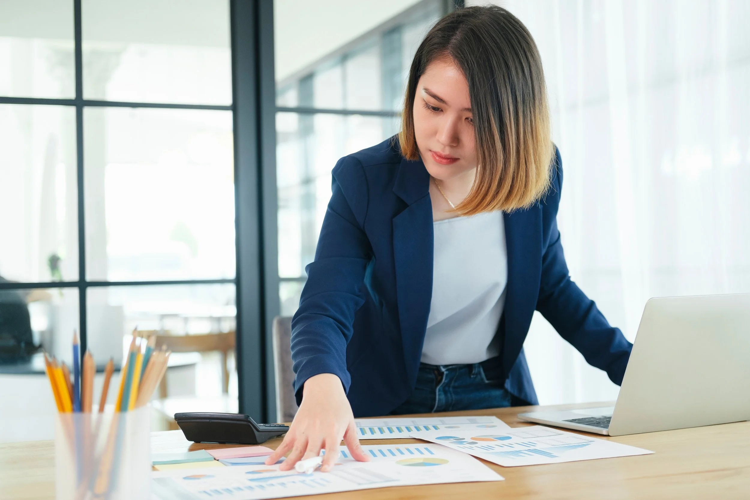 Businesswoman in a blue blazer working on documents with charts and graphs at a desk in an office.