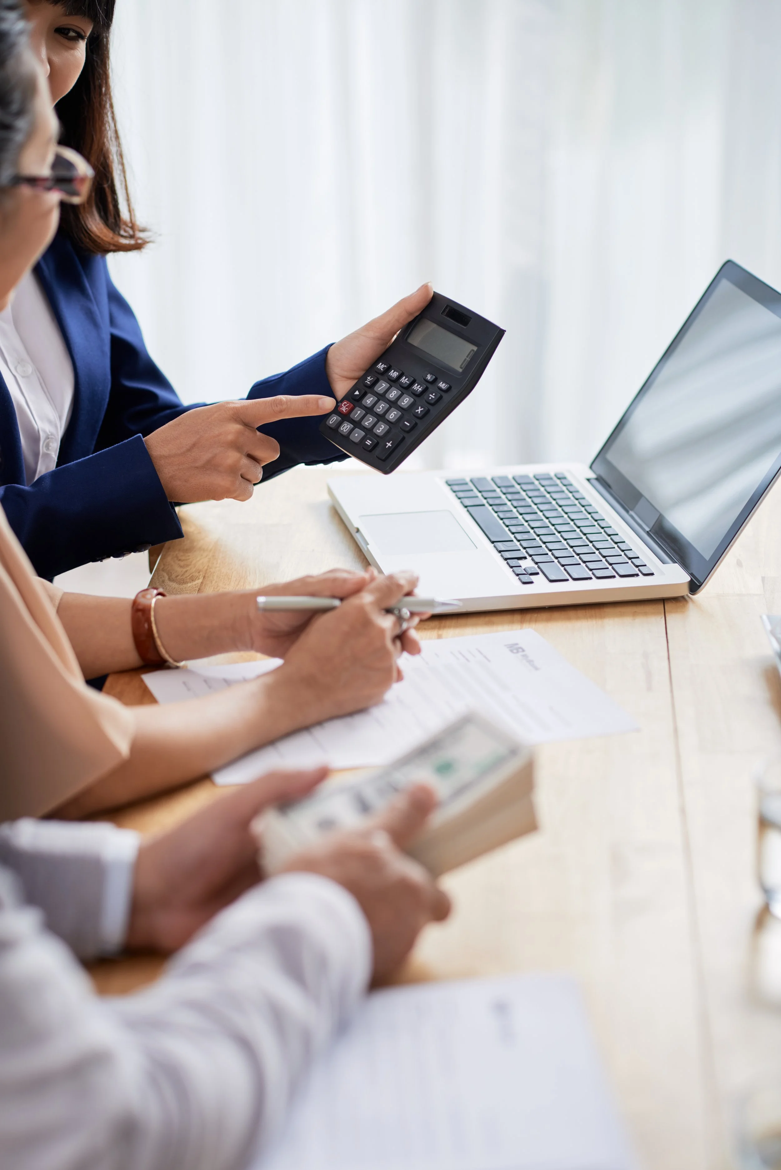 People working at a meeting with a laptop, calculator, cash, and documents on a wooden table.