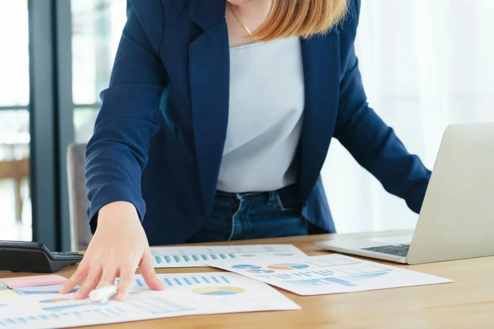 Businesswoman in a navy blazer and white top organizing documents with charts and graphs on a table alongside a laptop and a landline phone.