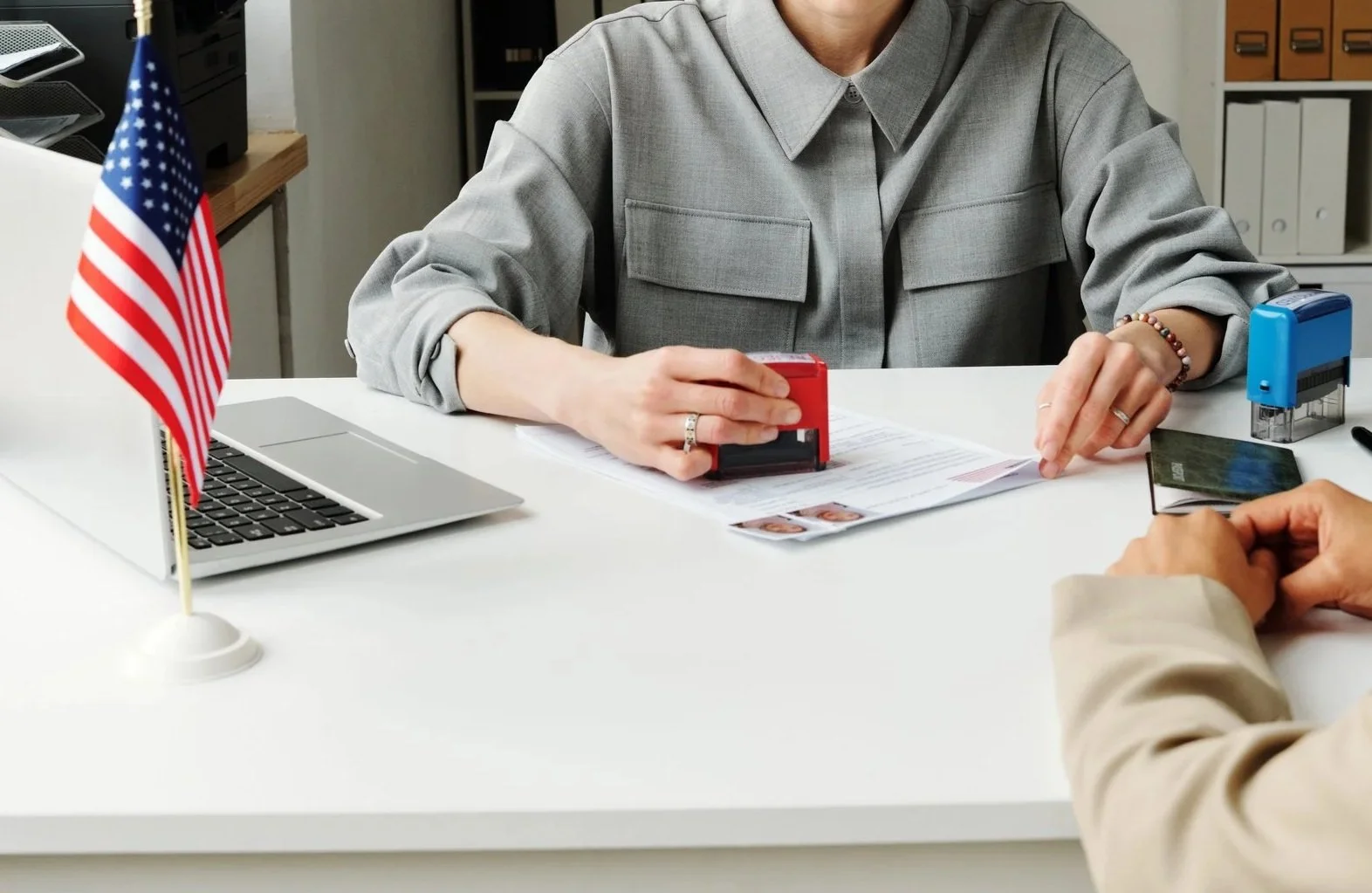 Person at an office desk with an American flag, a laptop, and stamped documents, receiving a passport from another person.