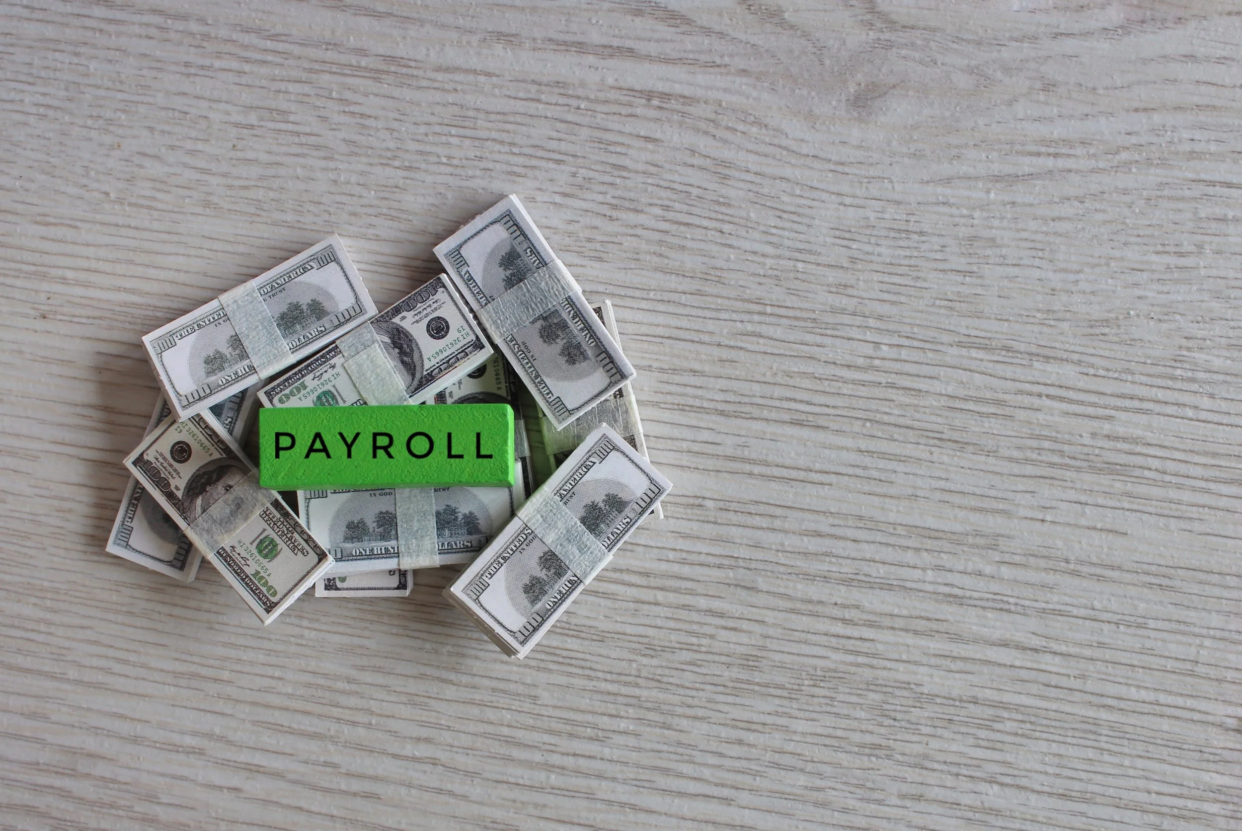 A pile of hundred-dollar bills with a green block labeled PAYROLL on top, placed on a light wood surface.