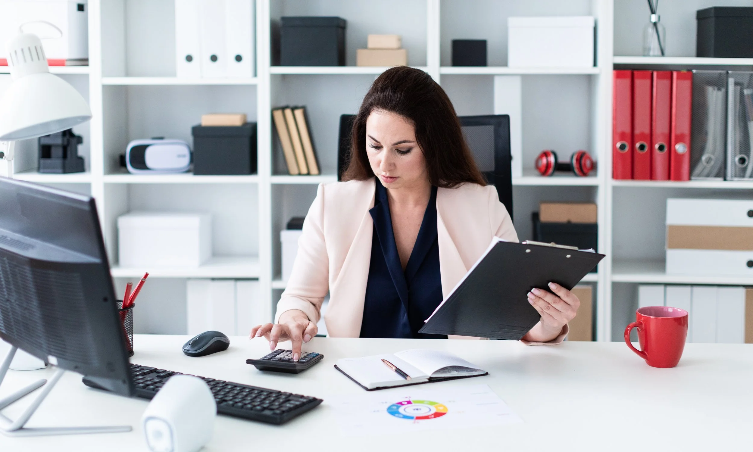 Woman working at a desk with a calculator, a computer, a keyboard, a mouse, a notebook, a diagram, and a red mug, in an office with white bookshelves in the background.