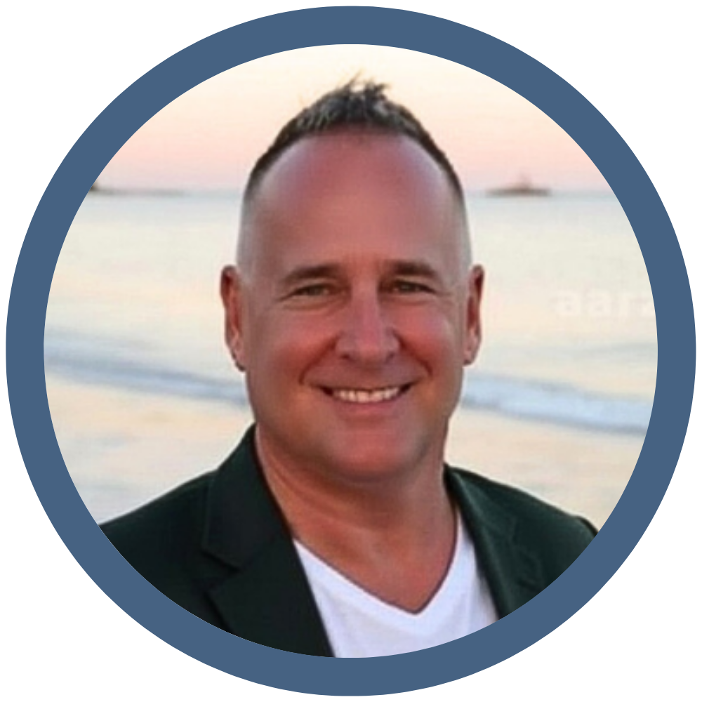 Smiling man with short hair and a dark blazer, standing on a beach with the ocean and sunset in the background.