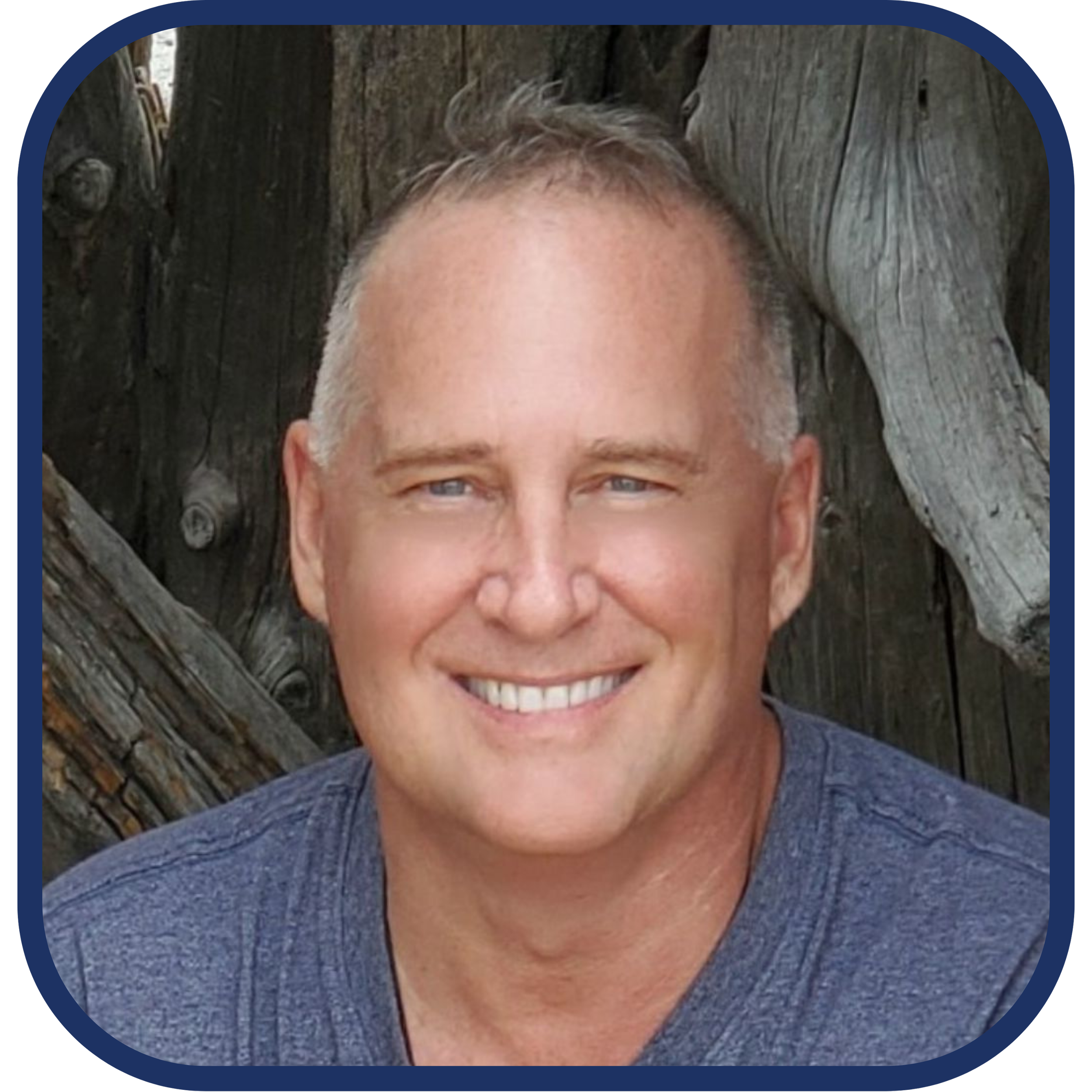 A smiling man with short gray hair wearing a blue shirt, standing outdoors in front of a wooden fence or wall.