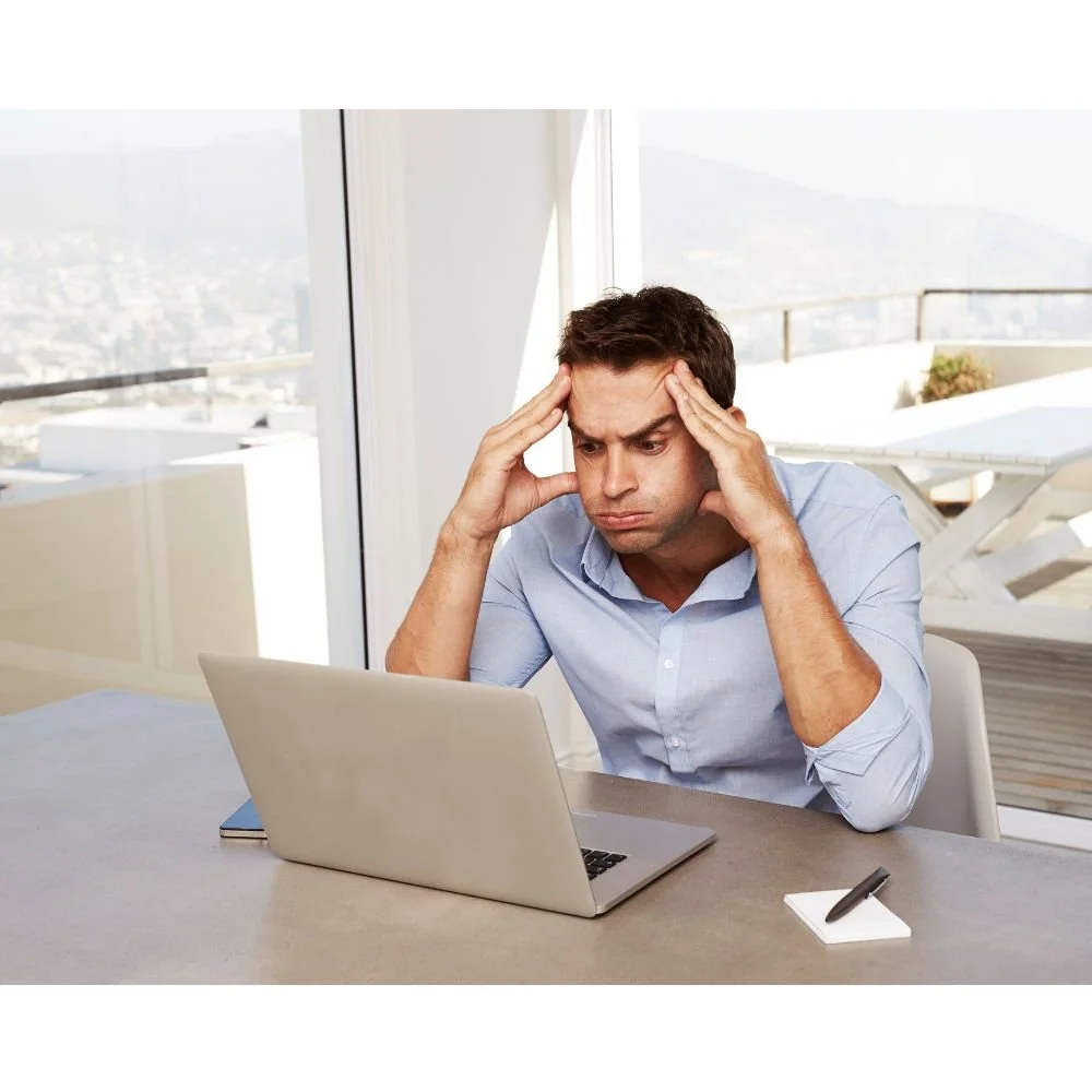 An anxious man sitting at a desk looking at a laptop with his hands on his head in a bright office with large windows.