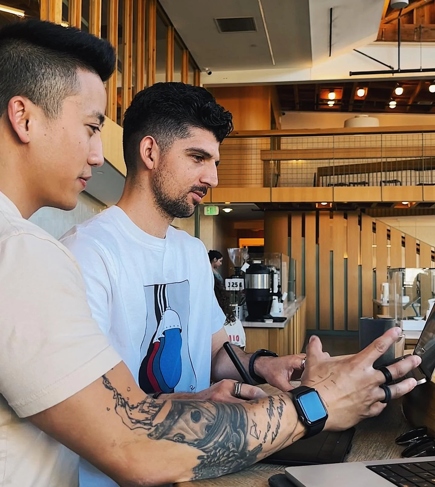 Two men sitting at a counter in a modern cafe, looking at and using a tablet. One man has tattoos on his arm and is wearing a smartwatch, the other has dark hair and a beard.