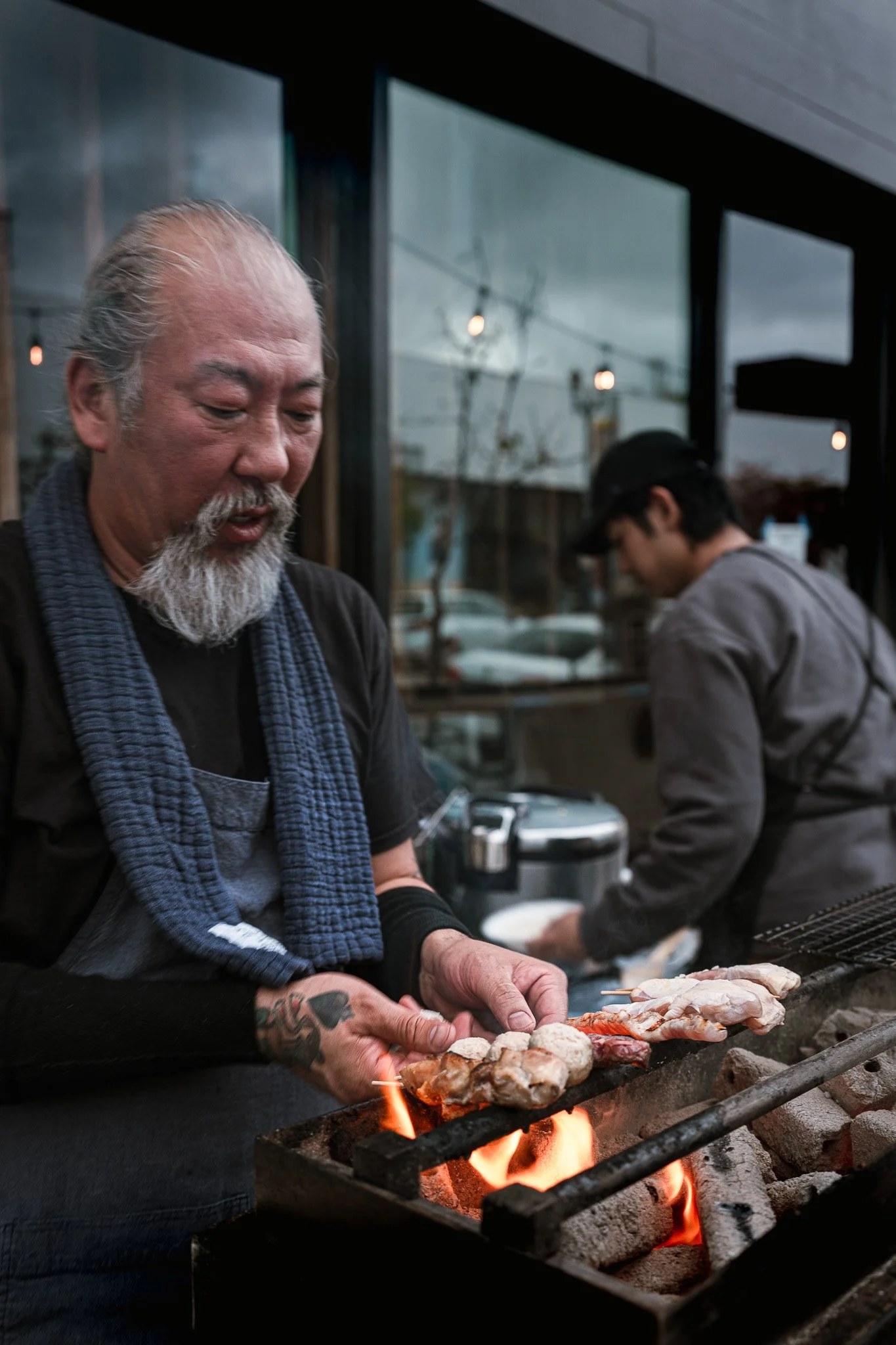 A photograph of an elderly man, named Yoya Takahasi, with a gray beard and tattoos on his forearm grilling skewered chicken over open flames outside a restaurant.