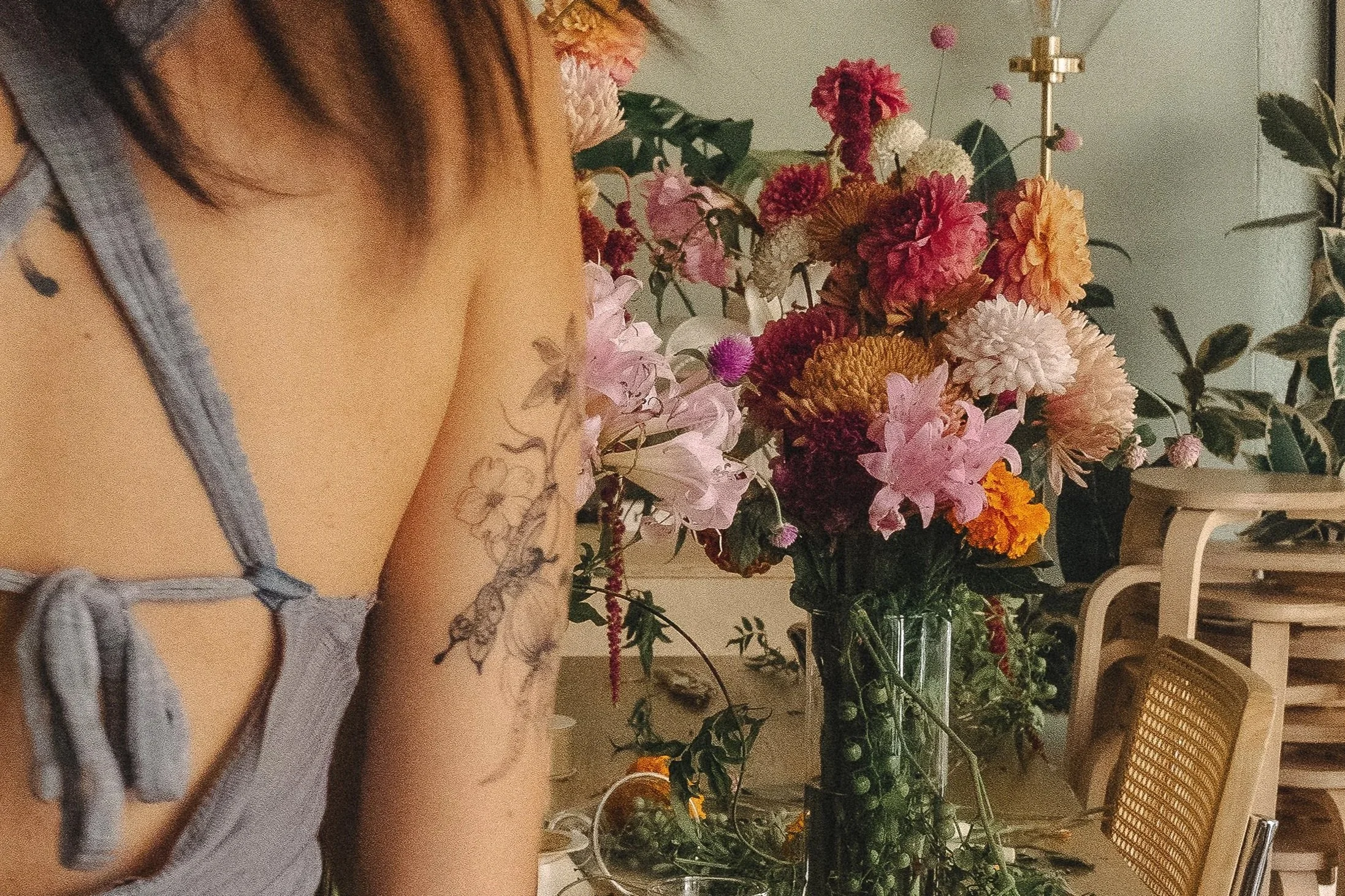 A woman with tattoos on her arm wearing a gray top, standing near a large bouquet of colorful flowers in a vase on a table inside a room.