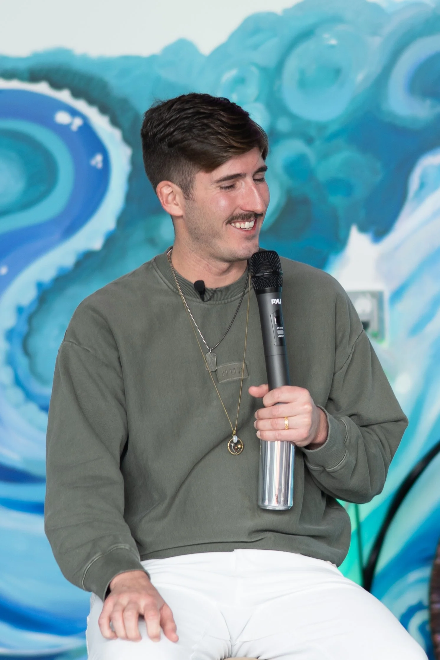 A young man with brown hair, mustache, wearing an olive green sweatshirt, white pants, and layered necklaces, sits holding a microphone, with a painted blue water-themed mural background.