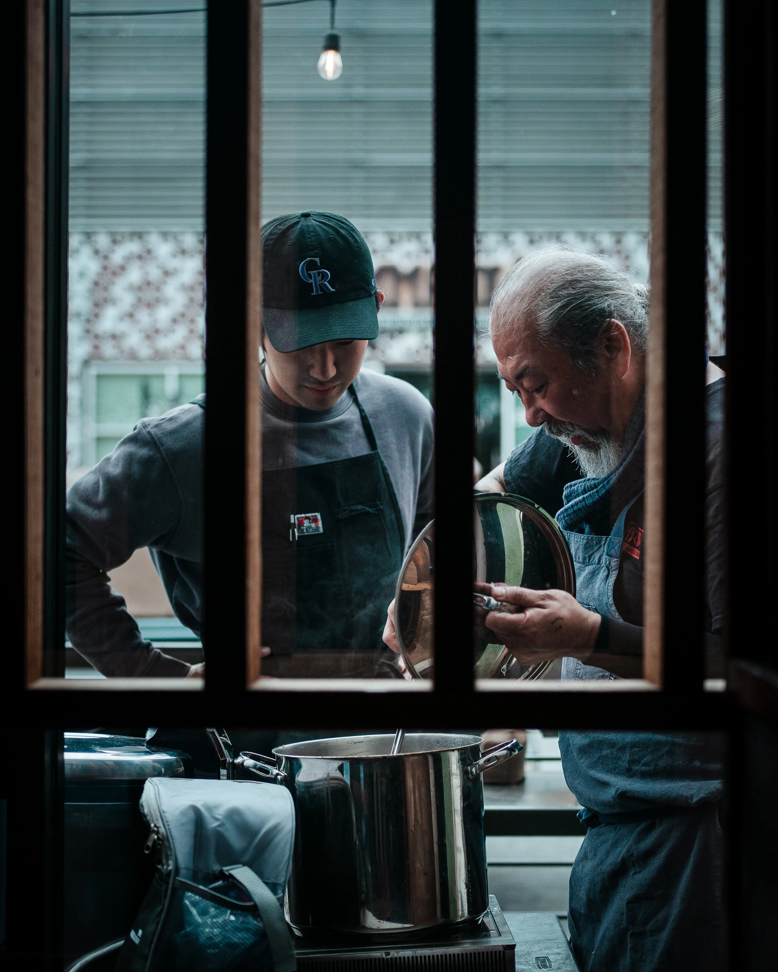 A photograph of Yoya Takashi--Two men cooking in a kitchen, seen through a window with black bars, one young wearing a baseball cap and the other older with a white beard, both wearing aprons, with a large pot on the stove.