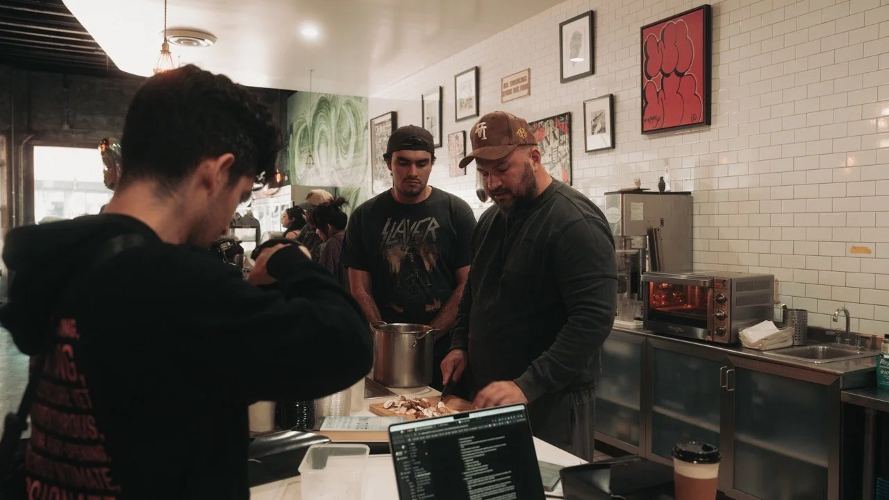 Group of people in a cafe kitchen, with a man in a baseball cap preparing food, while others watch, framed art on the white tiled wall.