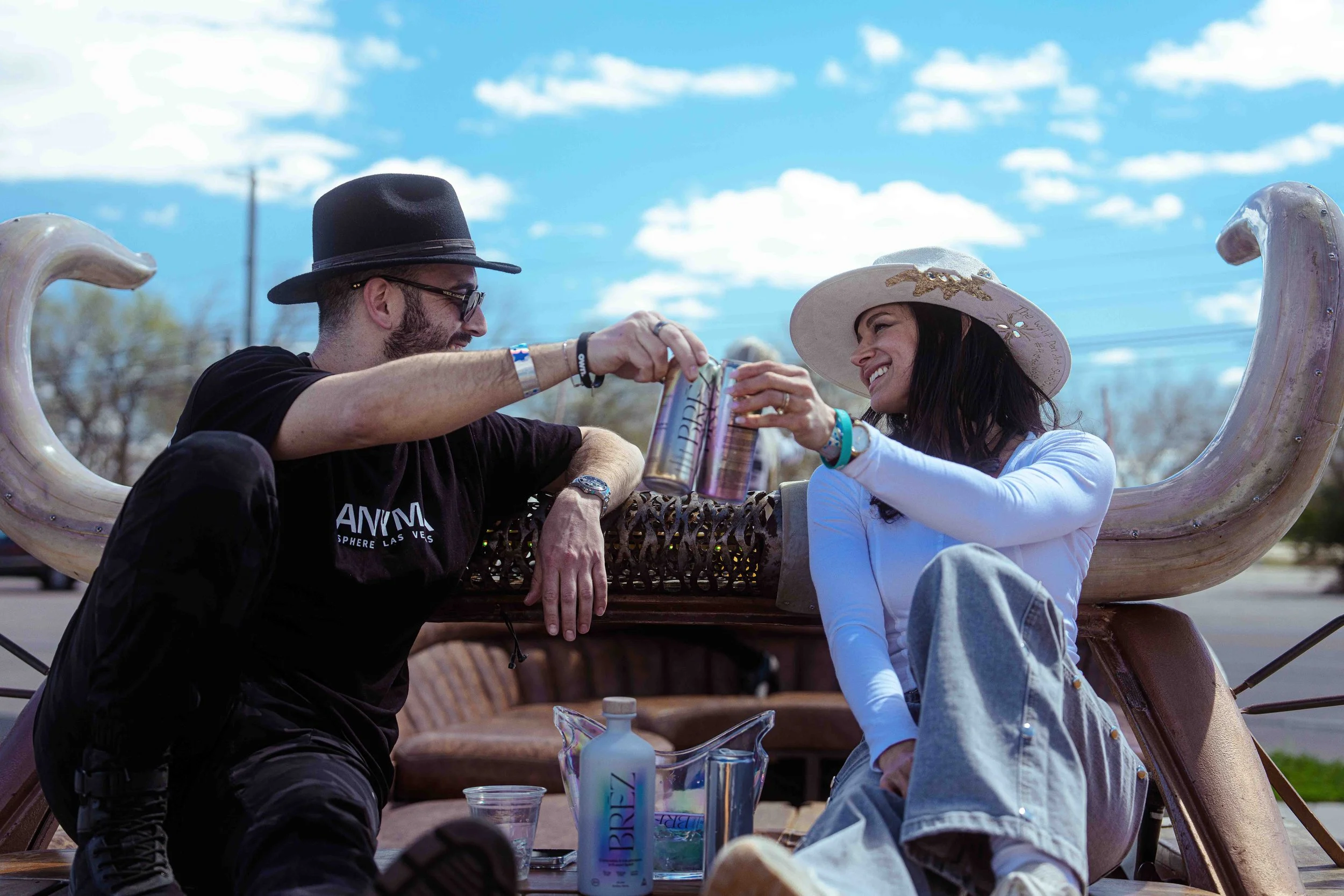 A man and a woman sitting on a decorated carriage or trailer, toasting with cans against a clear blue sky with scattered clouds, during an outdoor event or festival.