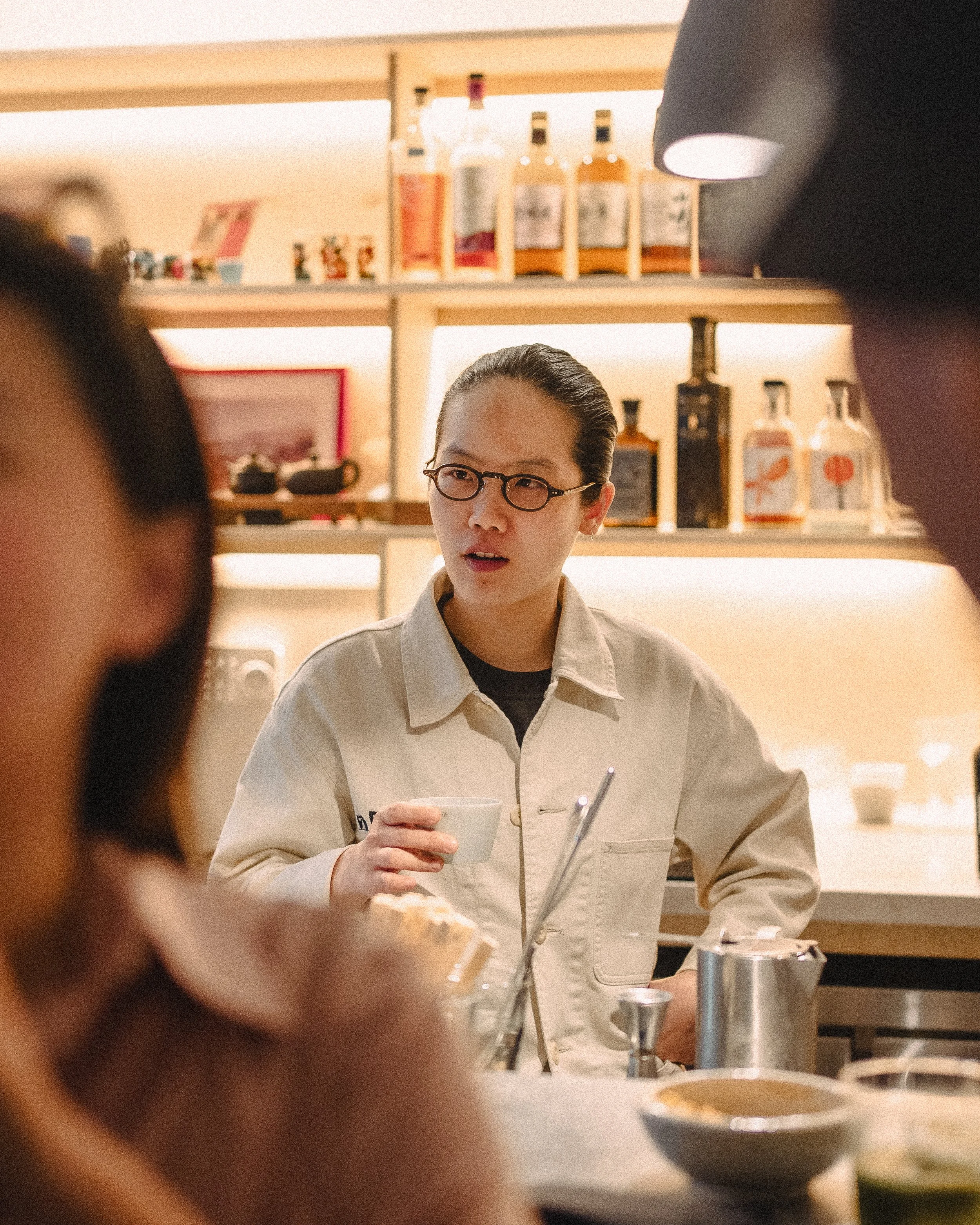 A person with glasses and a beige jacket holding a small bowl in a cafe or restaurant, with shelves of bottles and framed pictures behind them, and other people partially out of focus in the foreground.