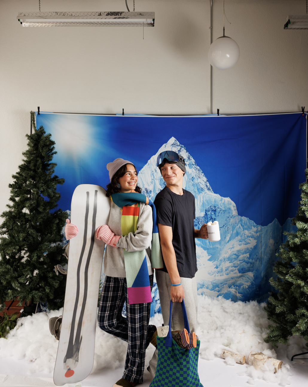 Two young people posing in a winter-themed setup with artificial snow, trees, and a mountain backdrop. One holds a snowboard, and the other holds a mug. They are dressed in winter clothes and smiling at each other.