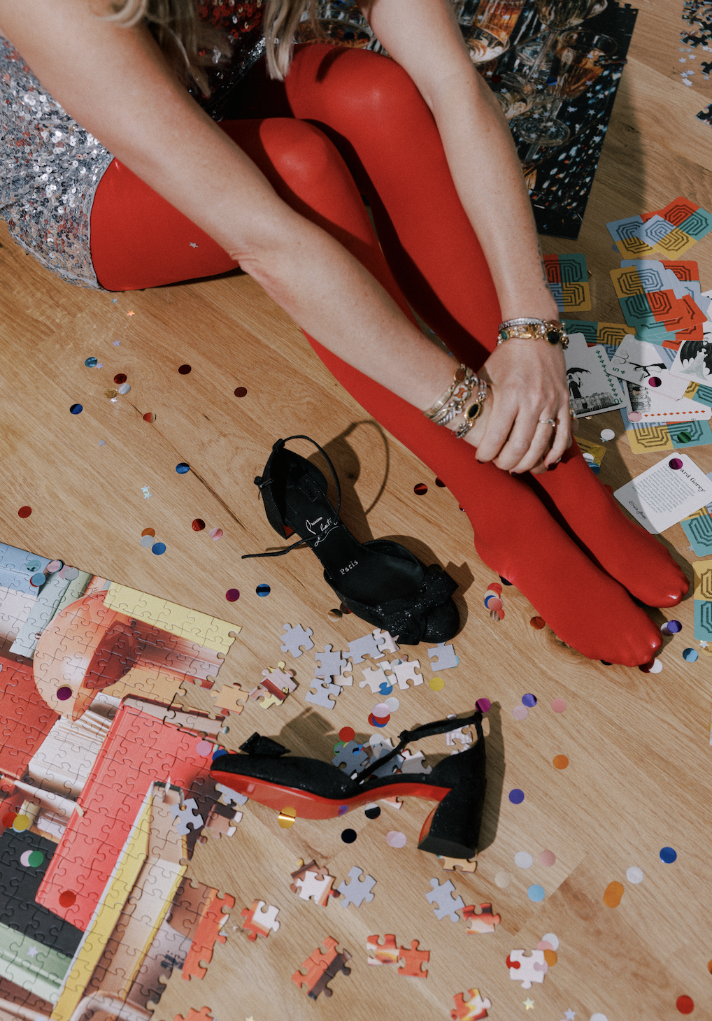 Person sitting on the floor during a celebration, putting on red tights, with scattered confetti, puzzle pieces, a pair of black shoes, and puzzle boxes around.