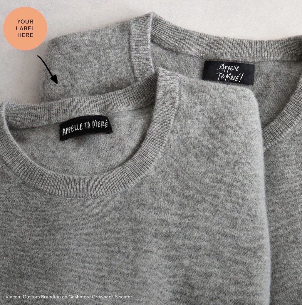 Two gray cashmere crewneck sweaters with black labels that read 'Appele Ta Mere!' and a customizable orange circle label labeled 'Your Label Here'.