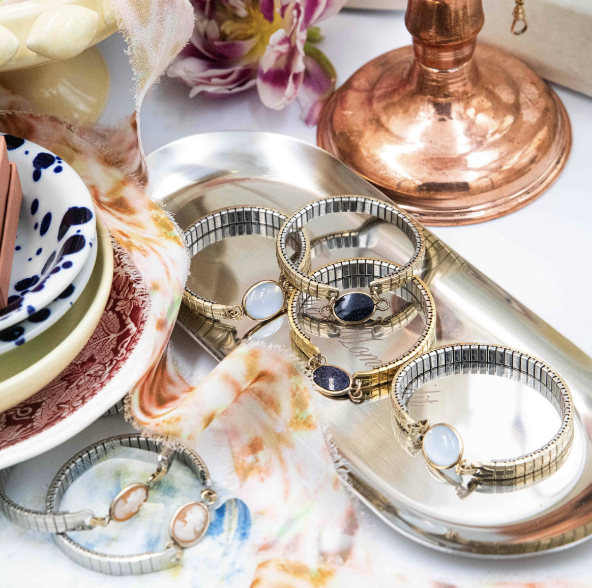 A collection of jewelry bracelets with large oval stones on a silver tray, surrounded by decorative bowls, floral fabric, and a copper candlestick holder.