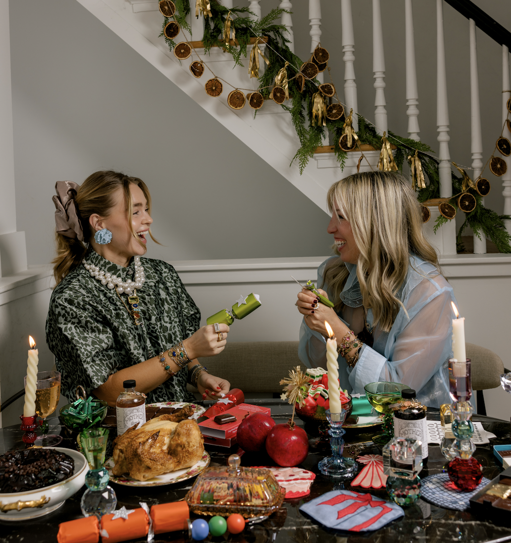 Two women celebrating at a festive gathering, surrounded by Christmas decorations and food, smiling and laughing while exchanging gifts.