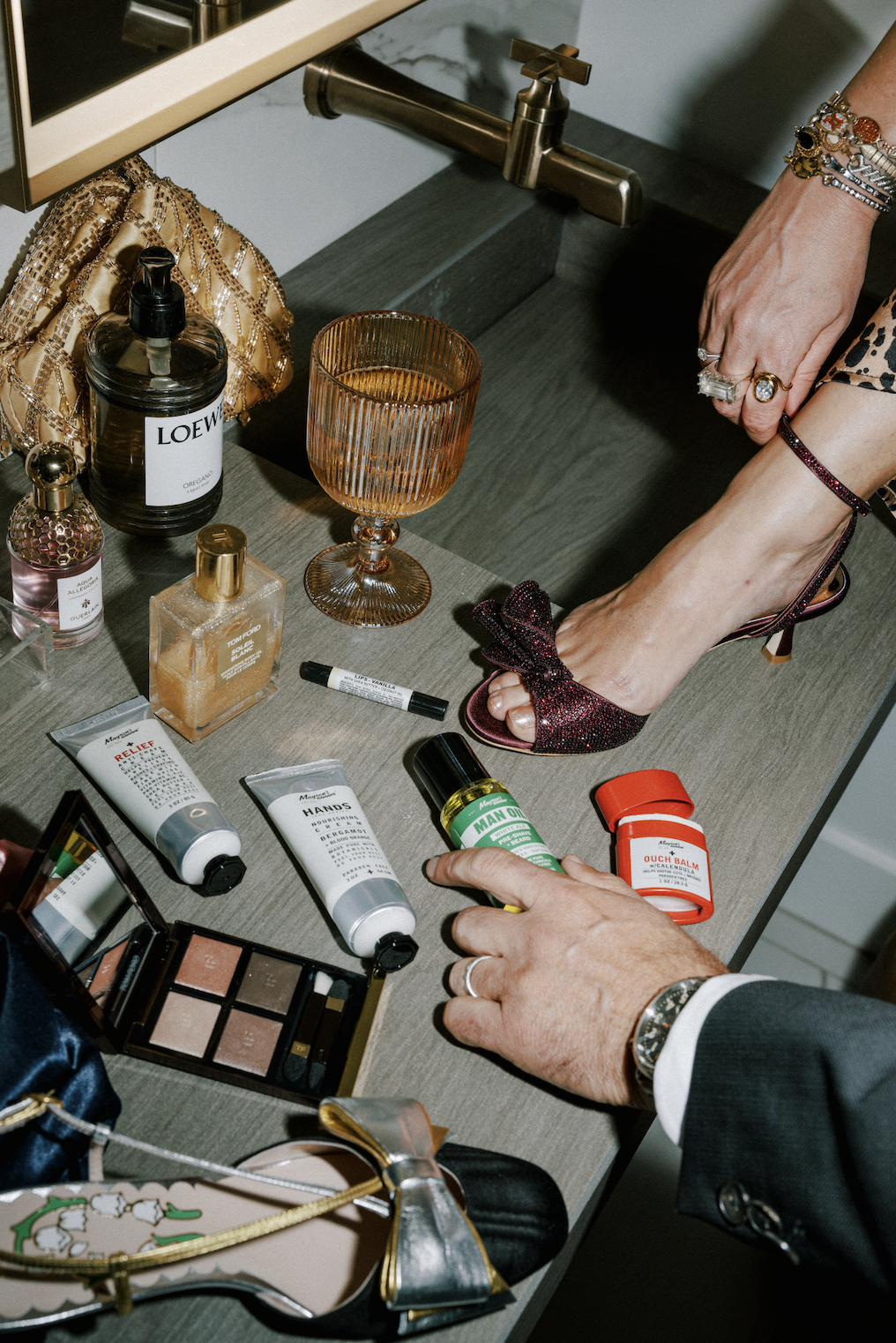 A woman is standing in a sink wearing high-heeled shoes, with various beauty products, perfume bottles, and makeup items on the countertop, and a man's hand reaching for a green ointment.