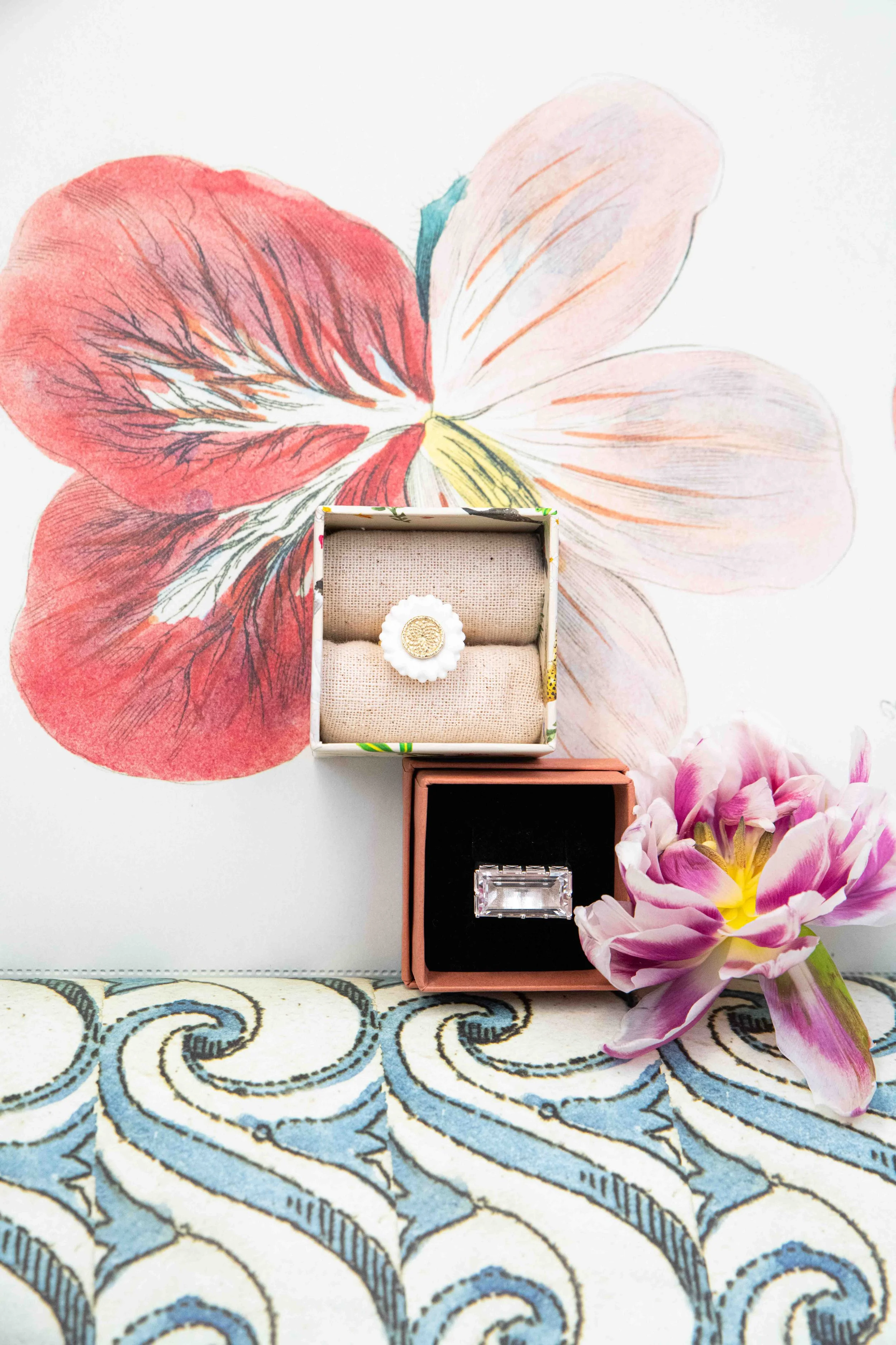 A decorative space with a large floral wallpaper in pastel colors, displaying a big pink and white flower. Two jewelry boxes, one with a ring and a watch, are placed in front of the wallpaper, with a bouquet of pink and white tulips on the right side.