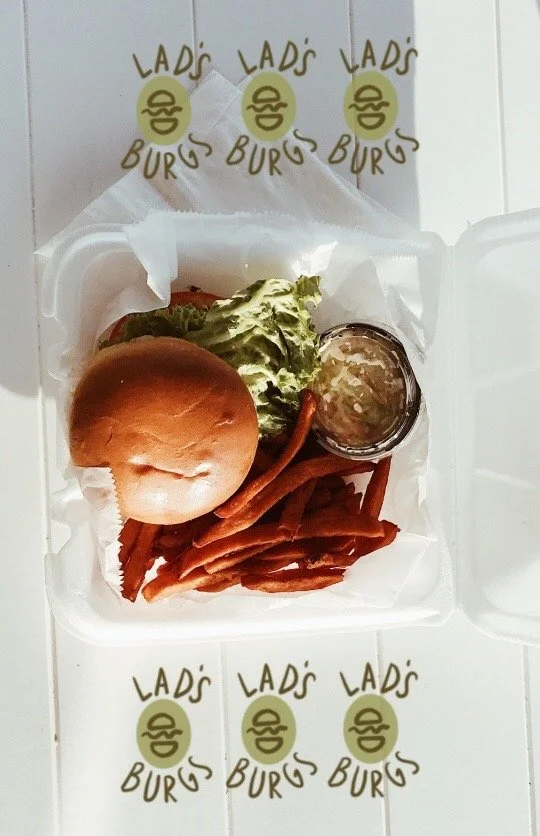A white takeout container with a burger bun, lettuce, and a small container of sauce, along with cooked bacon strips. The container is inside a box with repeated "Lad's Burgs" logos featuring a smiling burger face.