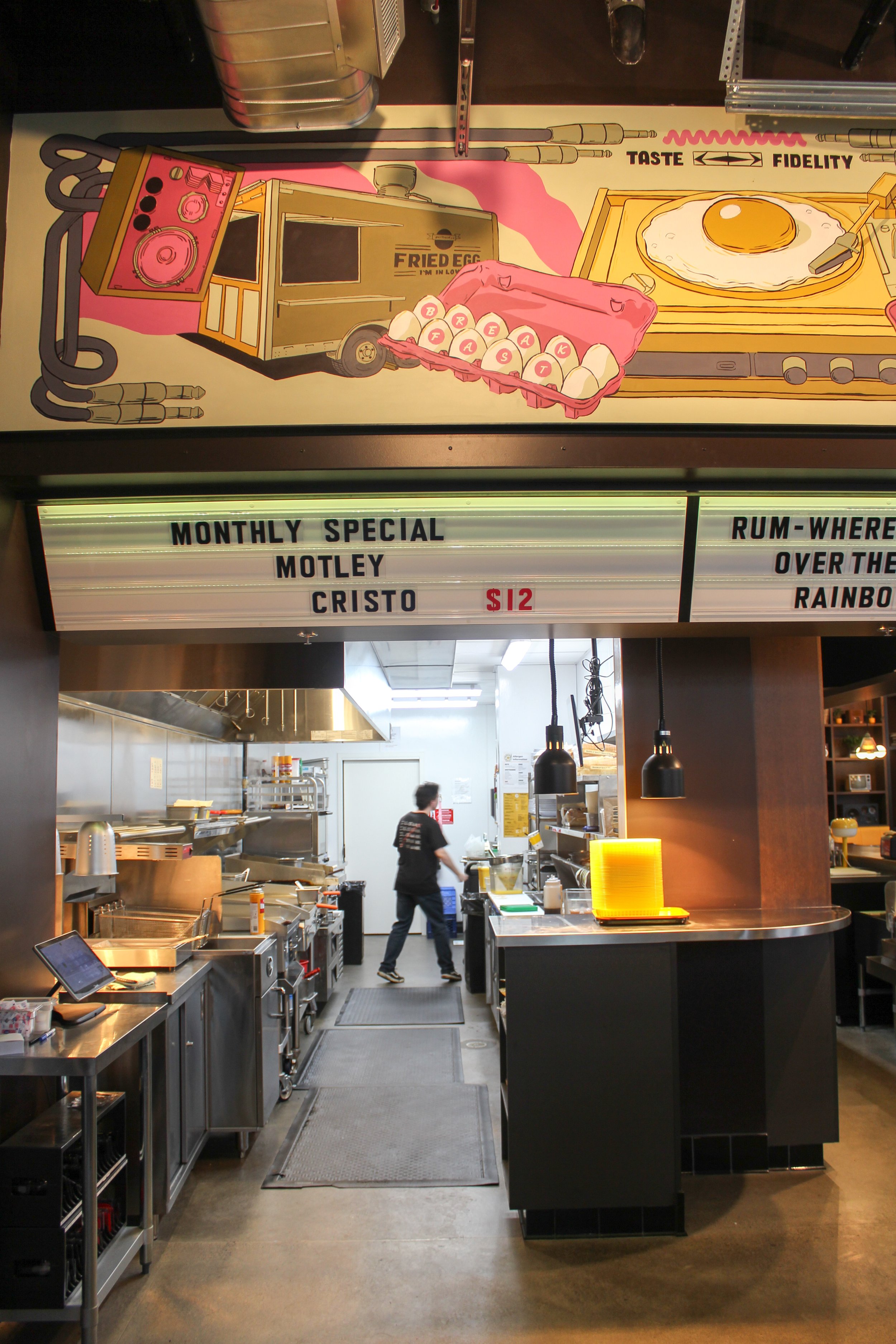 A restaurant kitchen with a person working, multiple stainless steel appliances, and a decorative sign above with an illustrated pink and yellow food truck, eggs, a fried egg on a stove, and a pink mixer. The sign features a menu with items and price