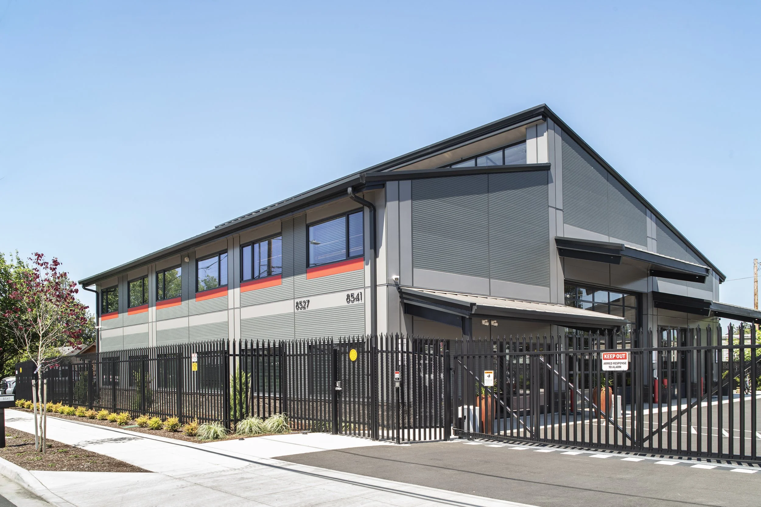 Modern two-story commercial or industrial building with metal siding, large windows, and a black fence surrounding a parking lot.