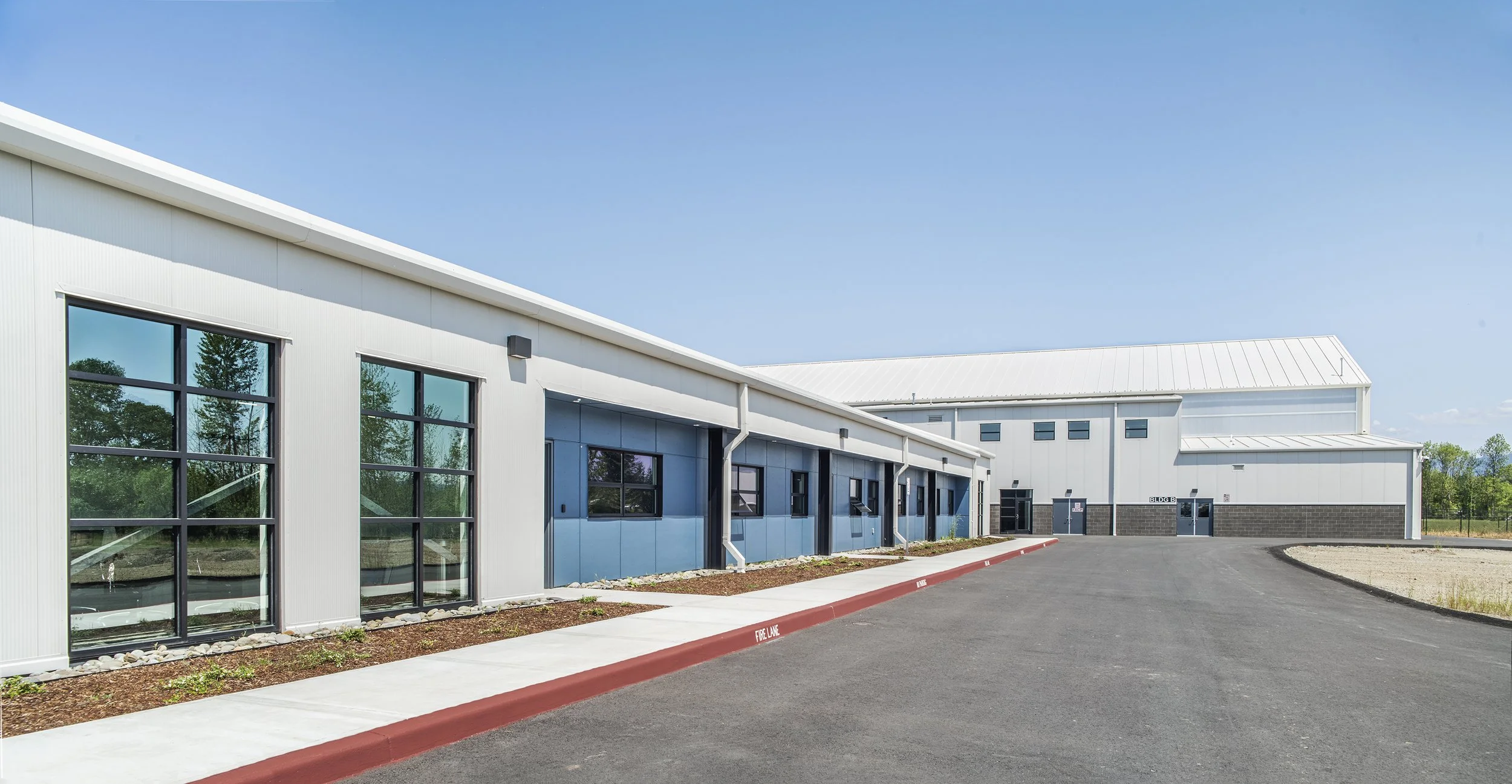Exterior view of a modern industrial building with metal siding, large windows, and a clean paved driveway under a clear blue sky.