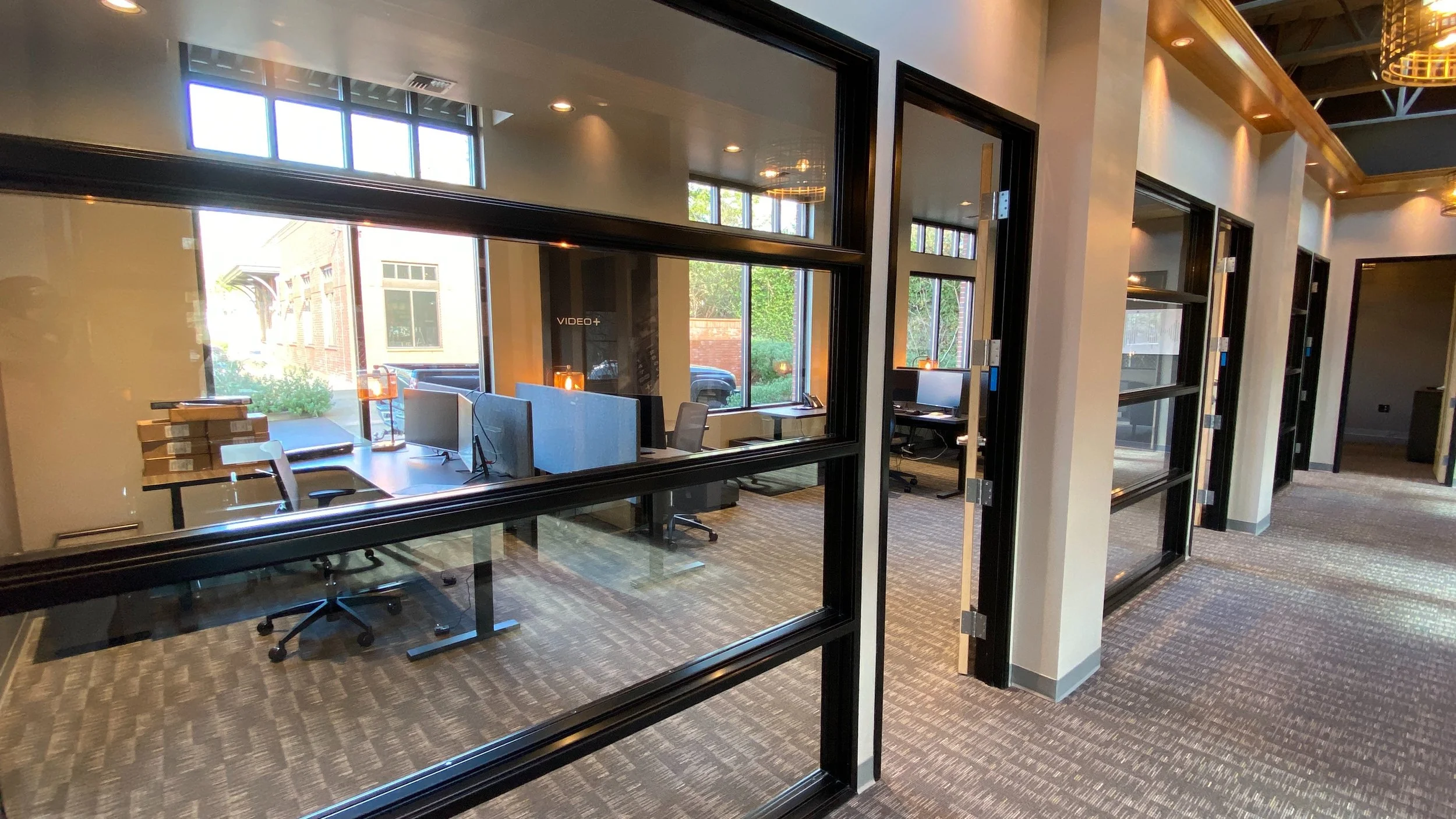 Empty office spaces with glass partitions, desks, chairs, and computers, illuminated by natural light from large windows.