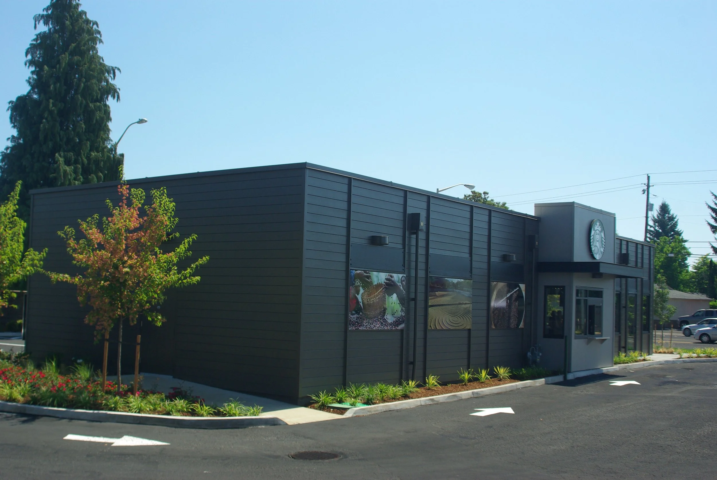 Exterior of a Starbucks coffee shop with black siding, a glass entrance door, and a Starbucks logo sign on a building with surrounding landscaping and parking lot.