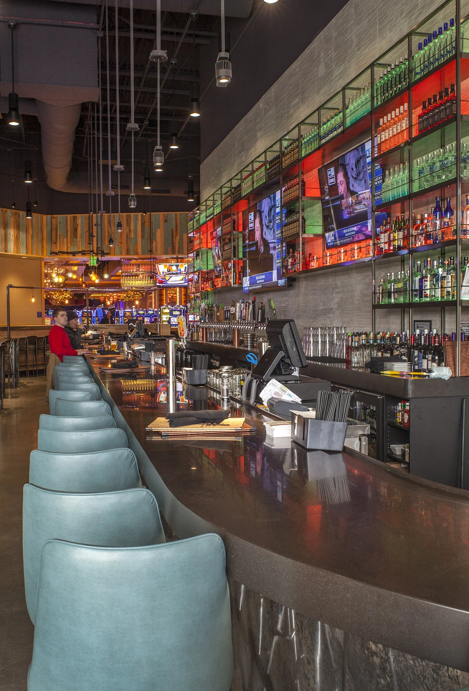 View of a modern bar in a restaurant or pub, with a curved dark countertop, teal cushioned bar stools, multiple TV screens showing news, shelves stocked with liquor bottles, and colorful lighting.