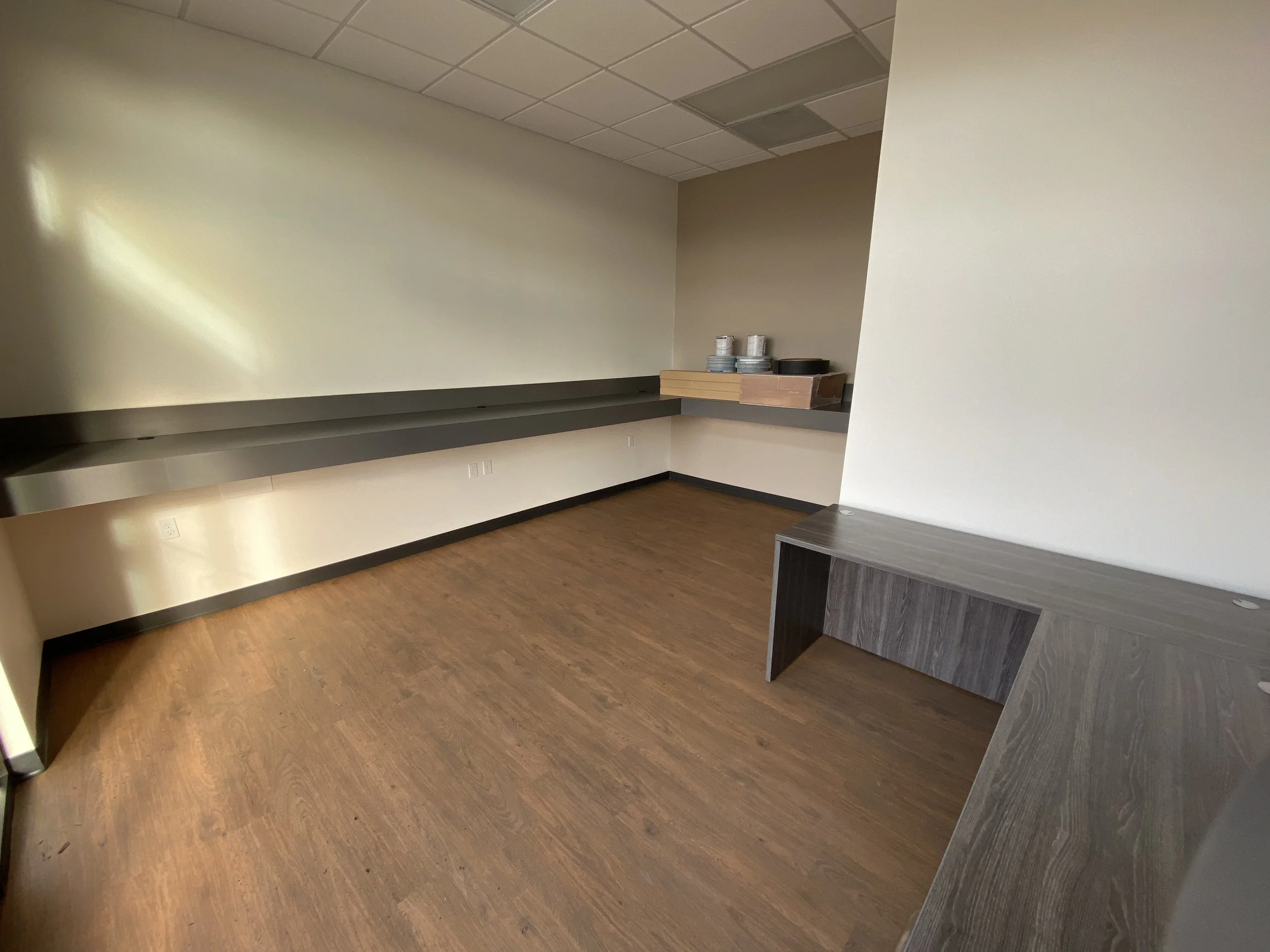 Empty room with wood flooring, beige walls, a long dark gray countertop along one wall, and a small dark wood desk in the corner. There are some plates and containers stacked on a small wall shelf.