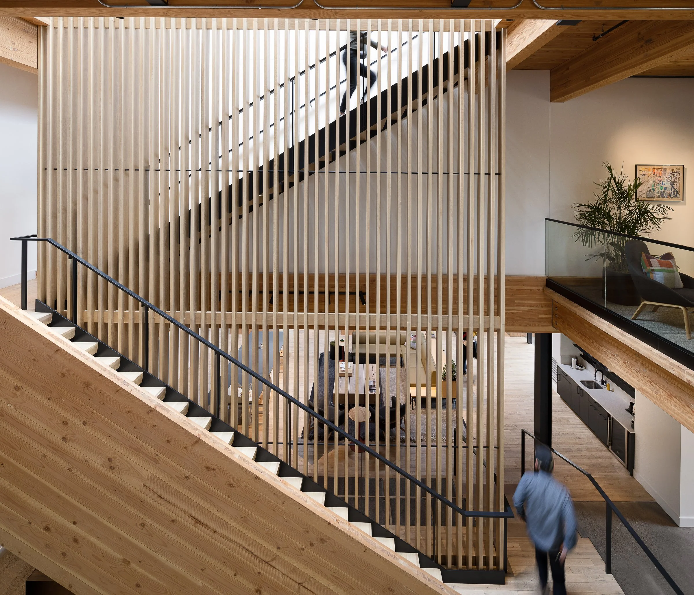 Interior of a modern building with a wooden staircase and a wooden slat divider. Two people are visible, one ascending the stairs and another descending. The space features a sitting area with a black chair, a colorful pillow, and a potted plant. A k