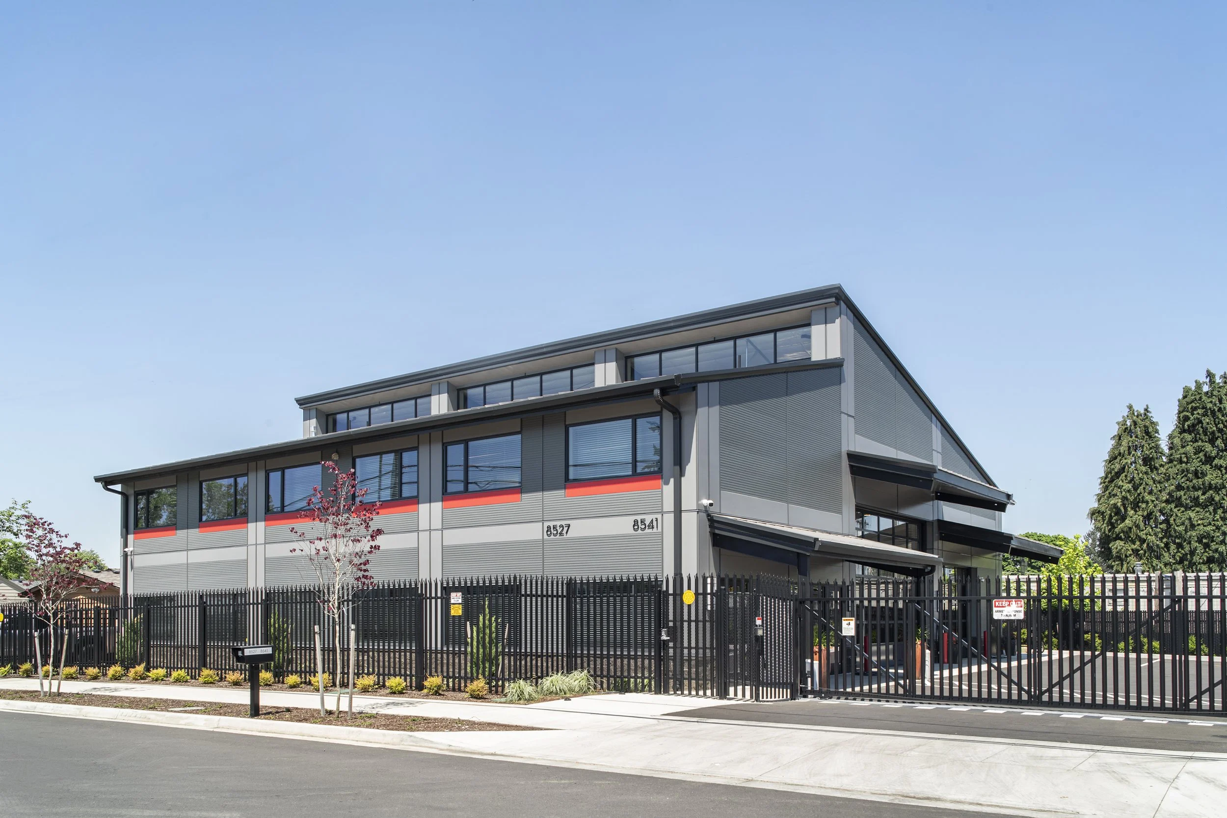 Modern commercial building with black fence, trees, and parking lot.