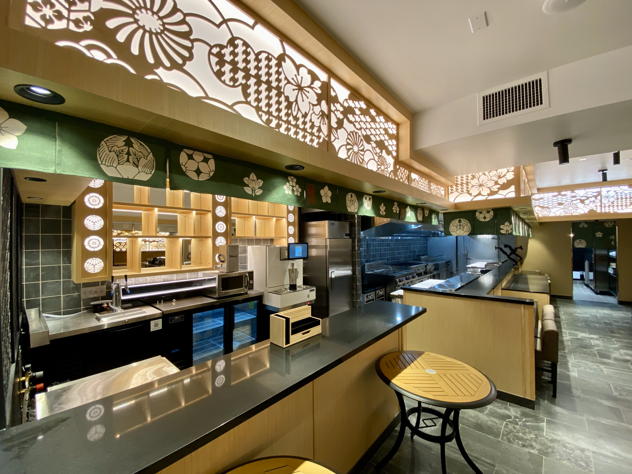 Interior of a modern Japanese-style restaurant kitchen with decorative paper cutouts of floral and geometric patterns on the upper panels, wooden shelving, a microwave, a refrigerator, and stainless steel cooking appliances, with a small round wooden