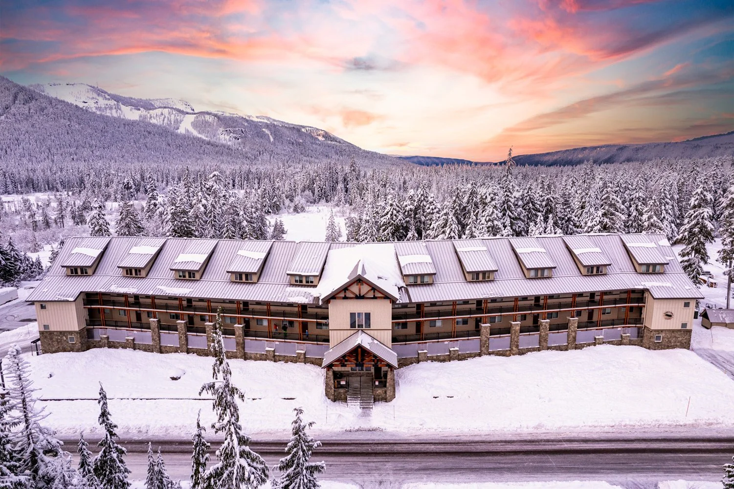 A snowy mountain landscape with a large hotel or lodge in the foreground, surrounded by snow-covered trees, under a colorful sky at sunset.