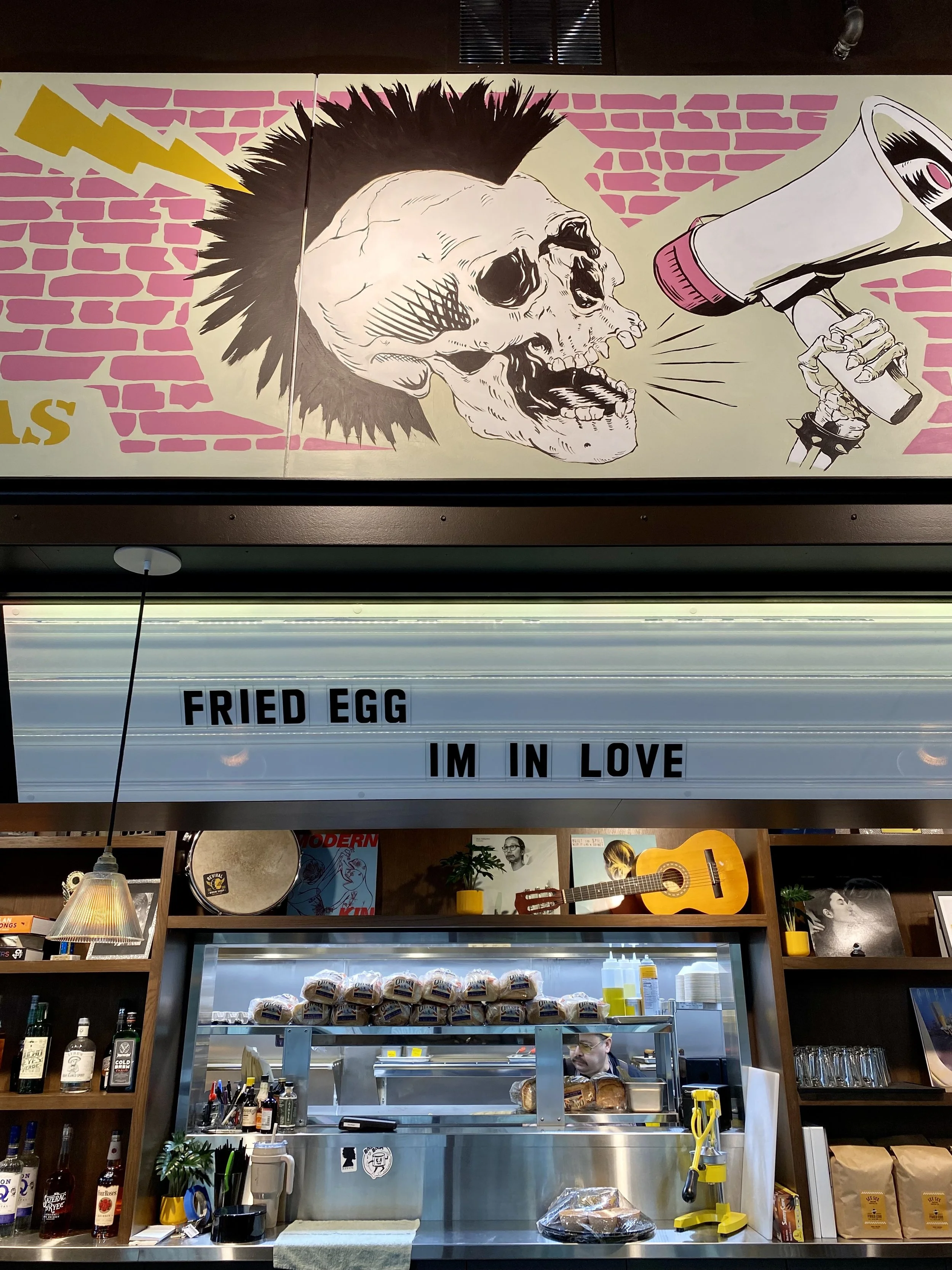 A mural of a skull with a mohawk, holding a megaphone, on a pink brick wall. Below, a sign reads "FRIED EGG I’M IN LOVE" in black letters. The cafe counter displays various items, with bottles, a guitar on the shelf, and a sign with the text.
