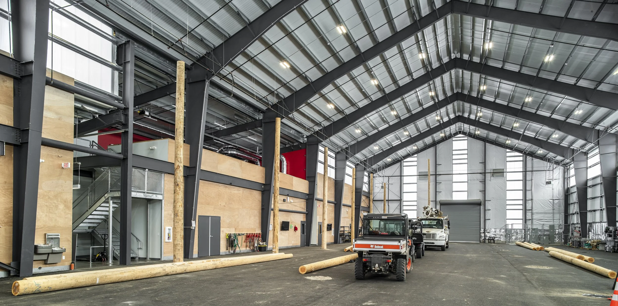 Interior of a large industrial warehouse with a high, arched metal roof, concrete floor, wooden beams, and construction equipment including a Bobcat loader and logs.