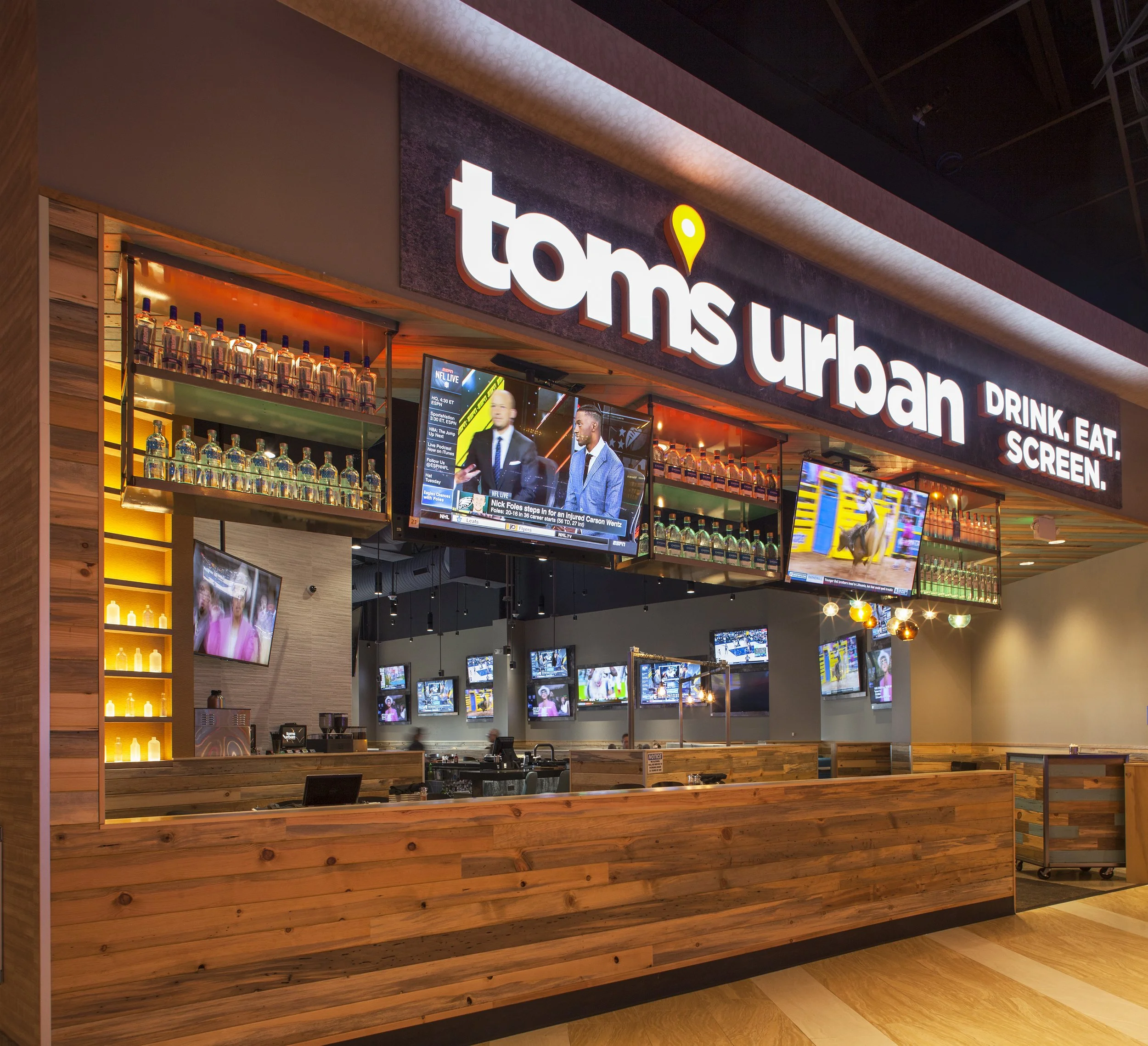 Interior of a restaurant named 'Toms Urban' with a bar area, multiple television screens displaying sports, and a modern decor with wood accents.
