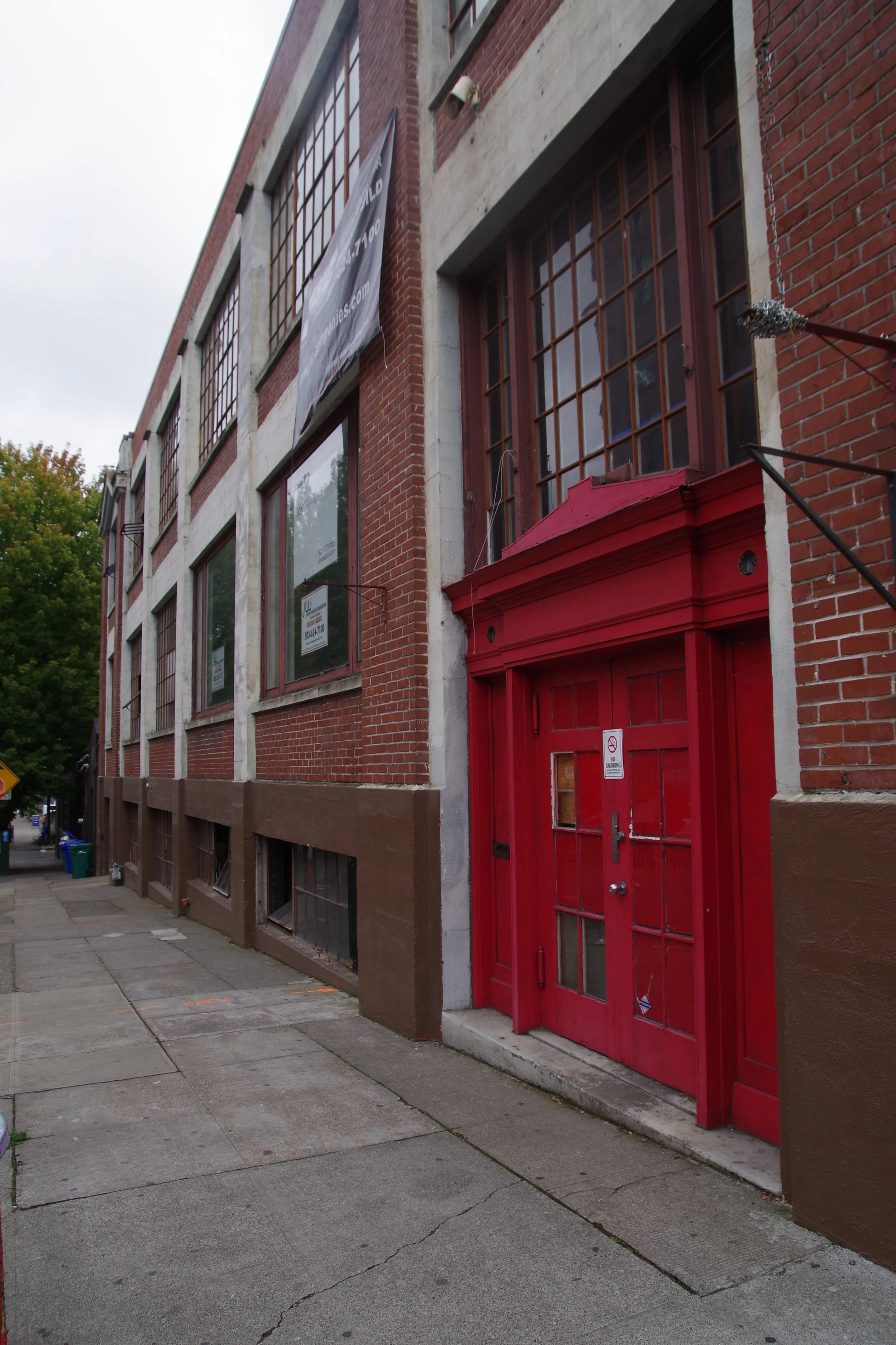 Red door entrance to a brick building with large windows and a transit shelter over the door, on a city sidewalk.
