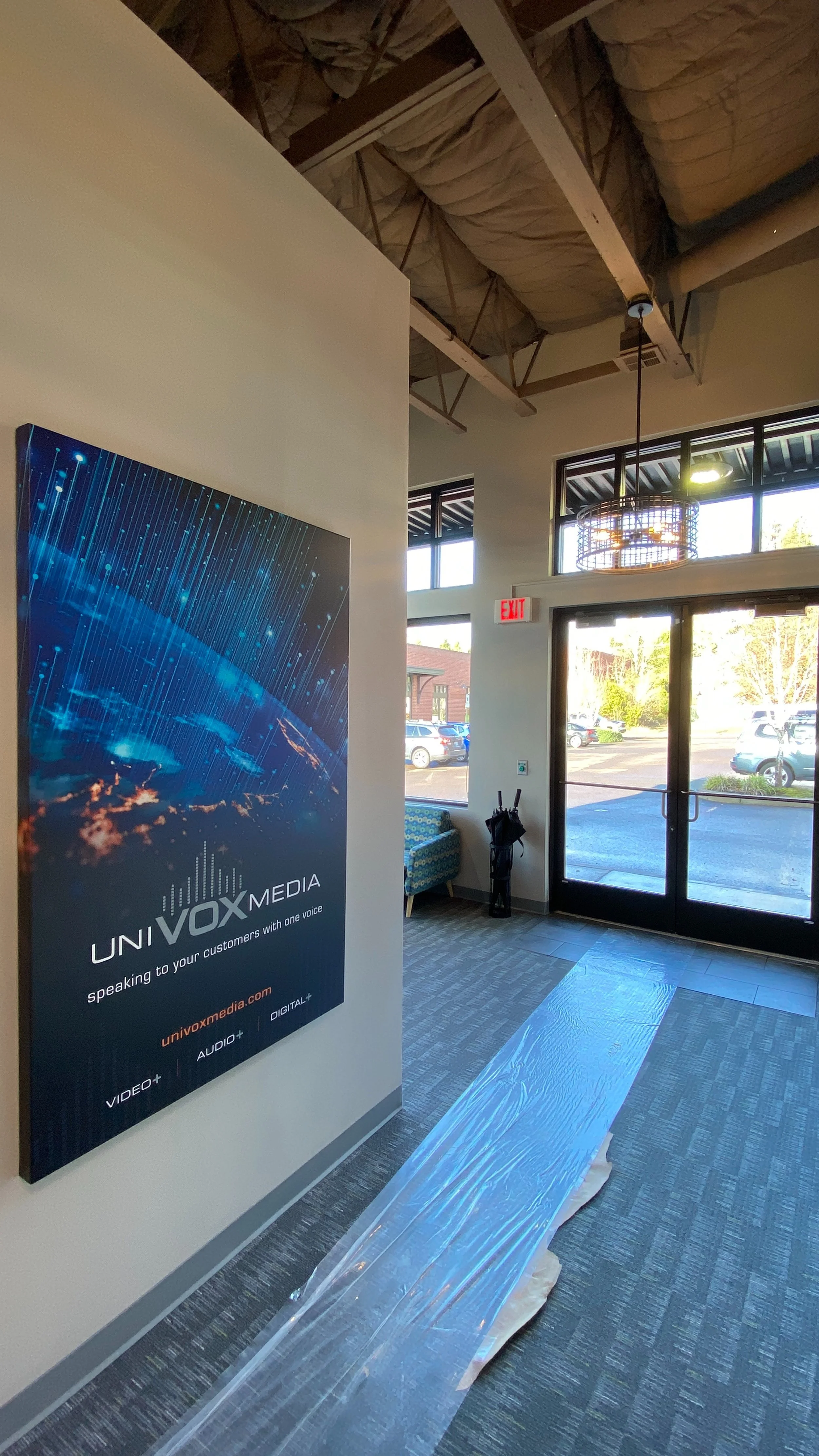 An interior view of a modern lobby area with a large digital media advertisement on the wall, showing a digital globe with data streams. There is a glass front door, a small seating area, and construction materials on the floor. The ceiling is unfini