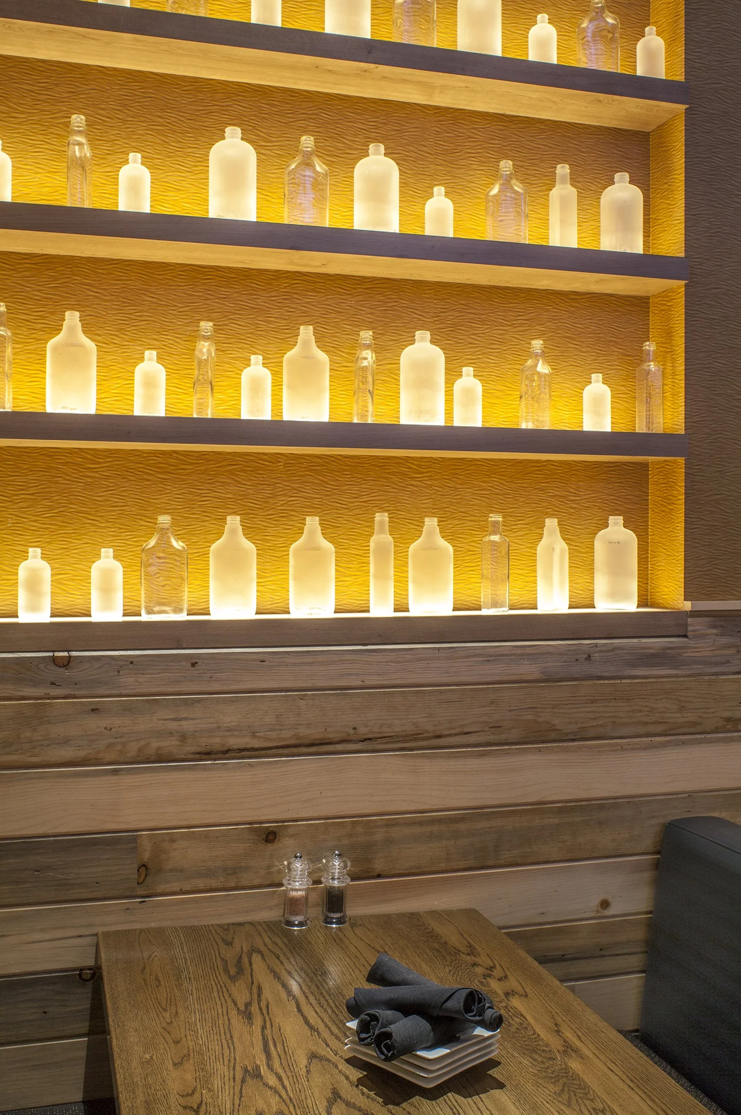 A wall with shelves displaying illuminated frosted and clear glass bottles, and a wooden table with cloth napkins, salt and pepper shakers in a restaurant or cafe setting.