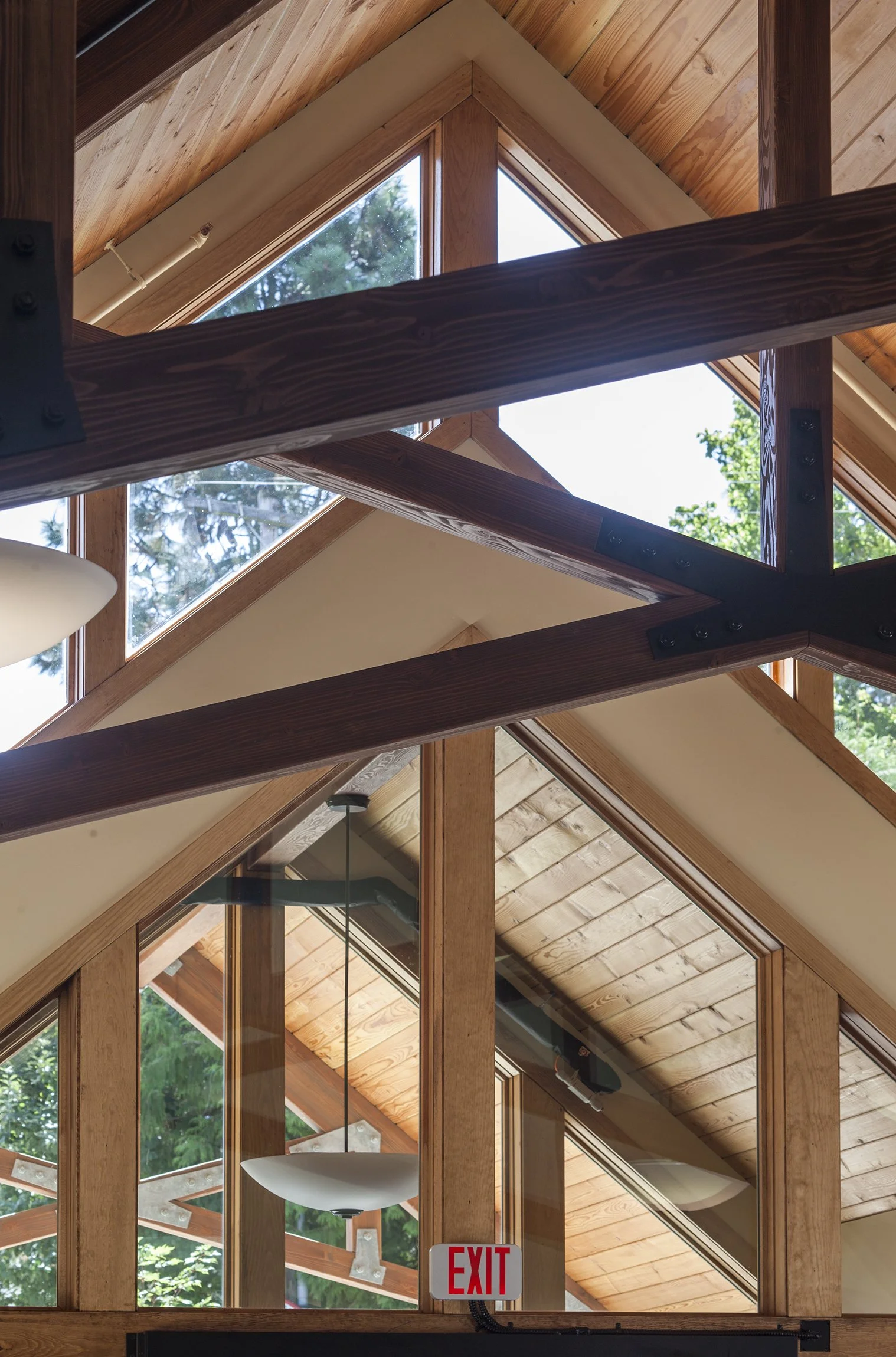 Interior view of a building with wooden beams, triangular windows, and pendant lights, with trees visible through the windows.